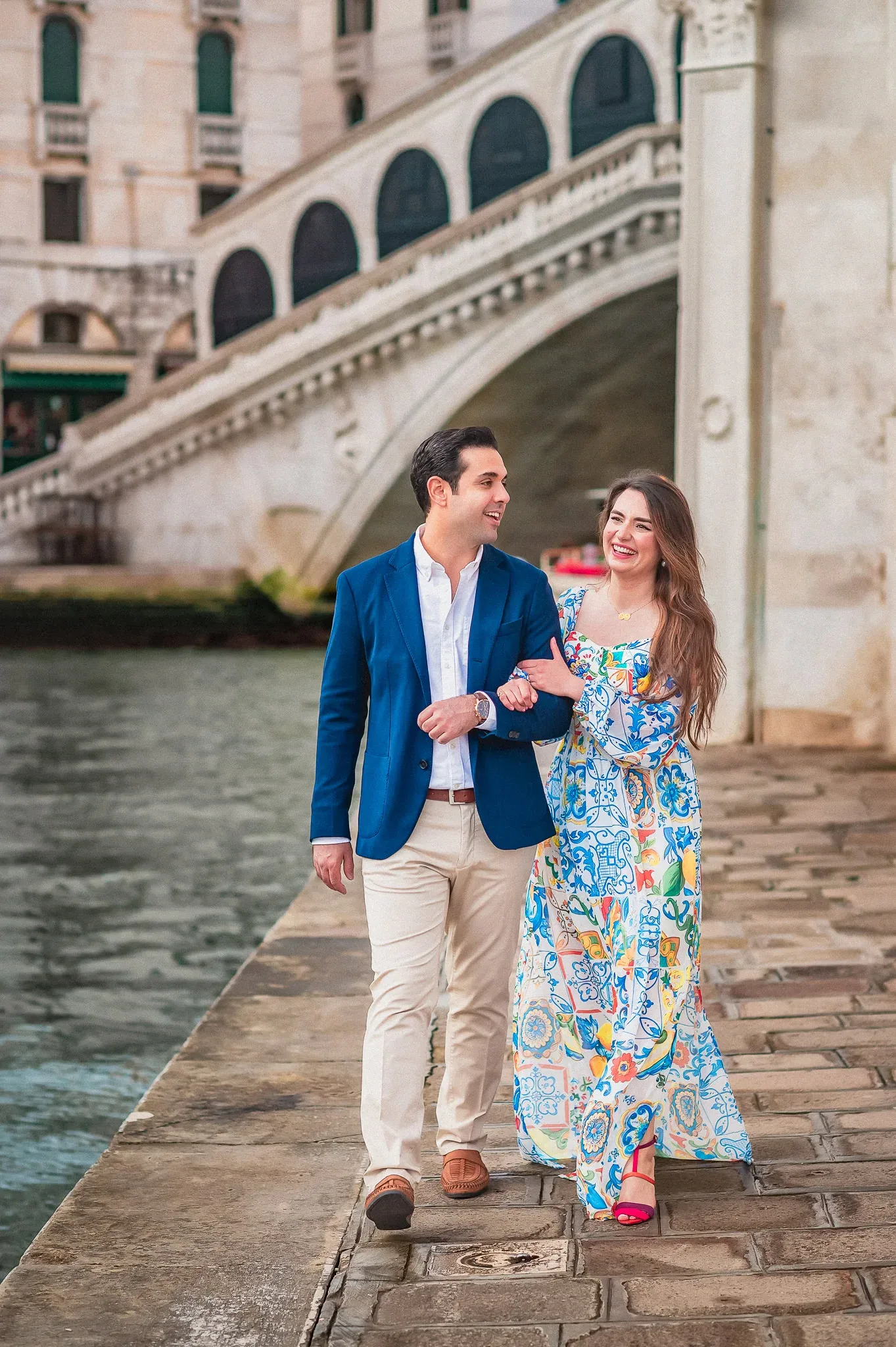 Romantic couple walking along Venice canal, engagement photo under historic bridge, colorful attire, joyful smiles, romantic Venice scenery.