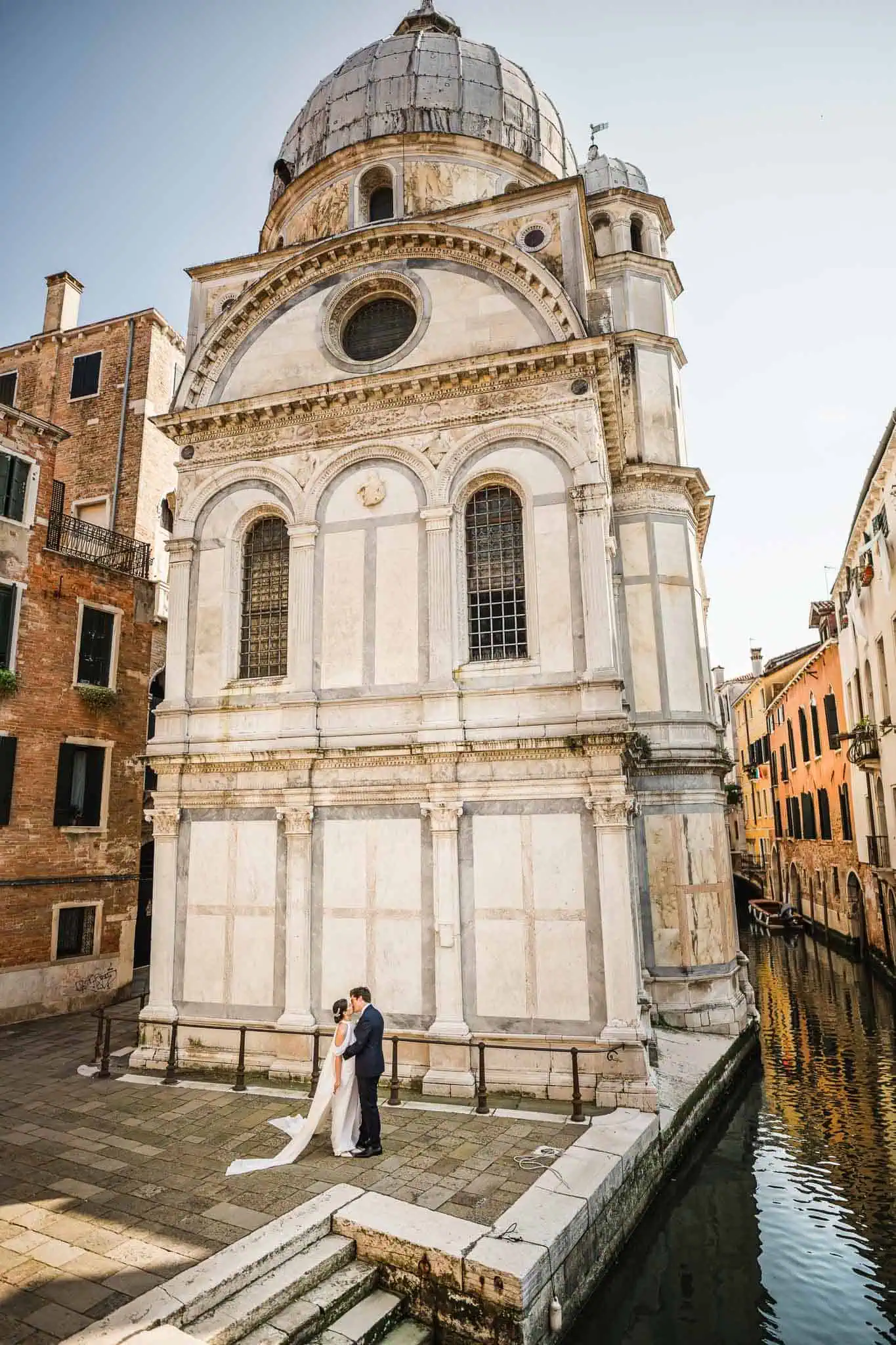 Romantic couple in wedding attire by Venice church, historic canal backdrop, daytime.