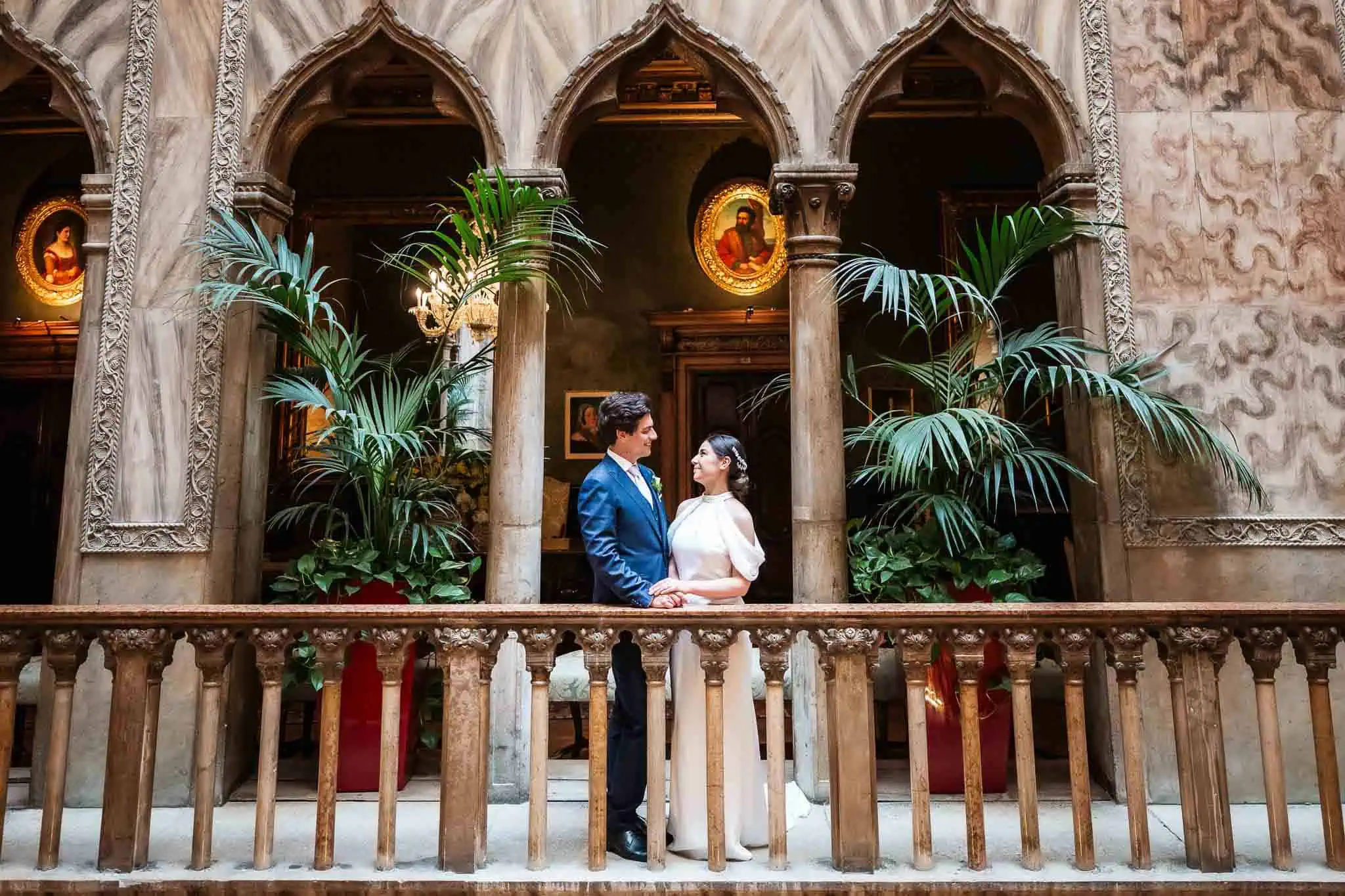 Elegant couple in wedding attire exchanging vows inside historic Venetian building with lush green plants.
