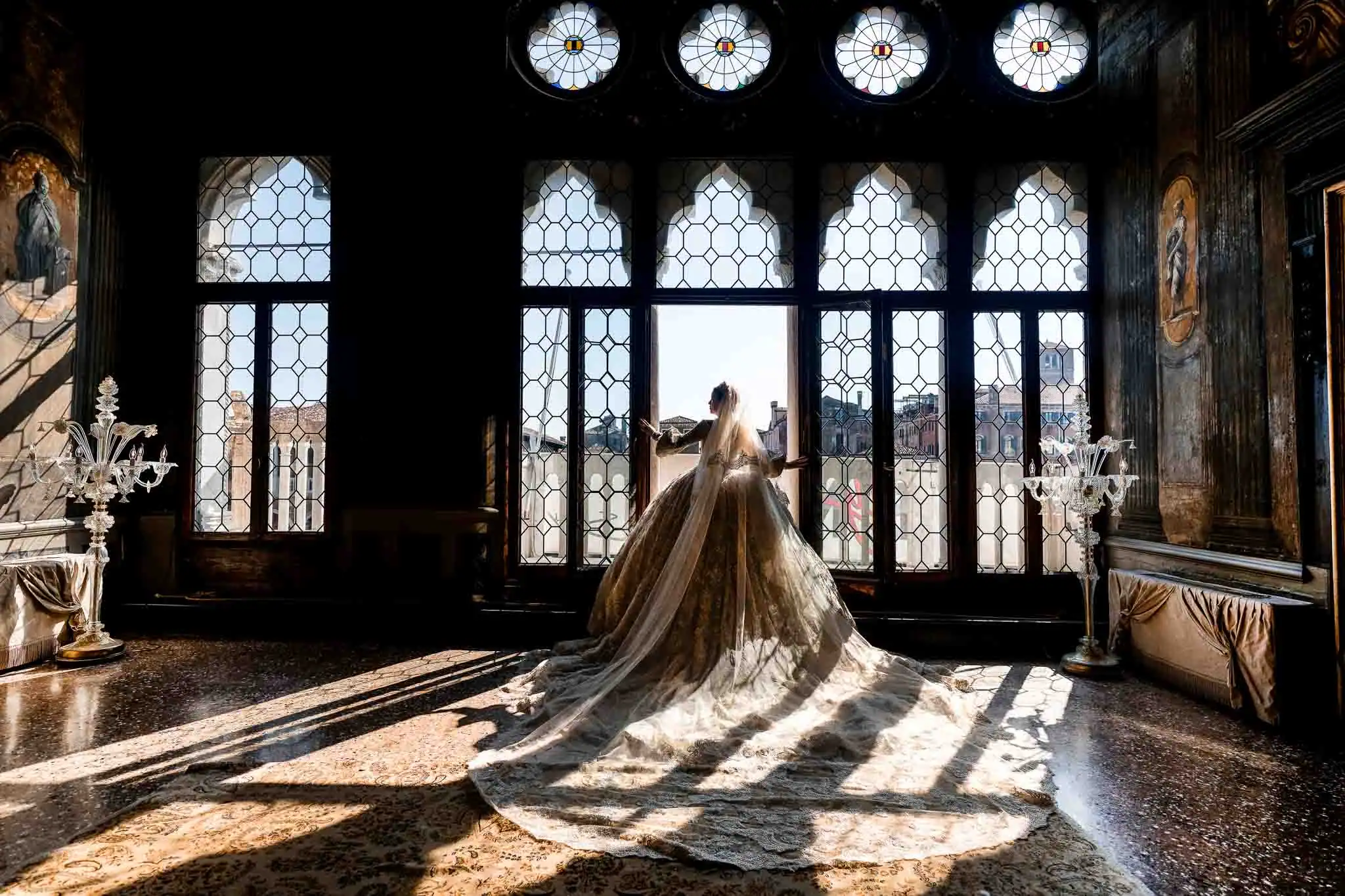 Elegant bride in vintage gown with sunlight streaming through historic Venetian windows.