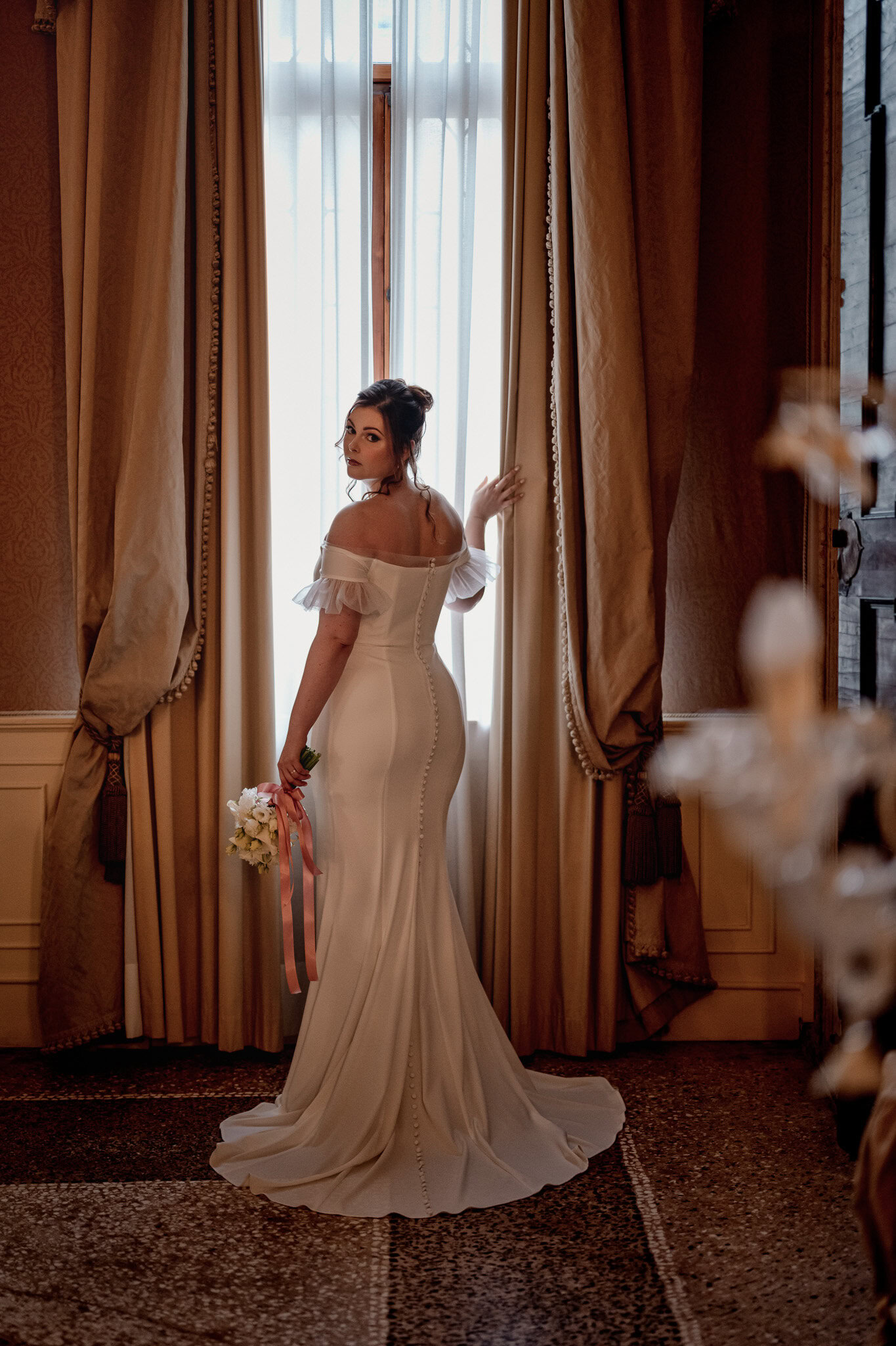 A woman in an elegant off-shoulder dress standing by a large window with golden curtains a.