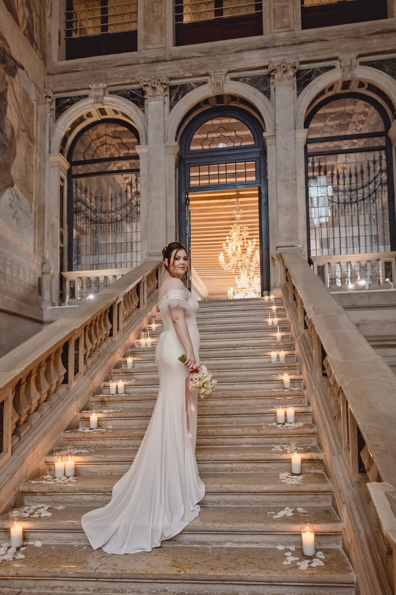 A woman in a white gown standing on a grand staircase at Ca’ Sagredo Palace in Venice.