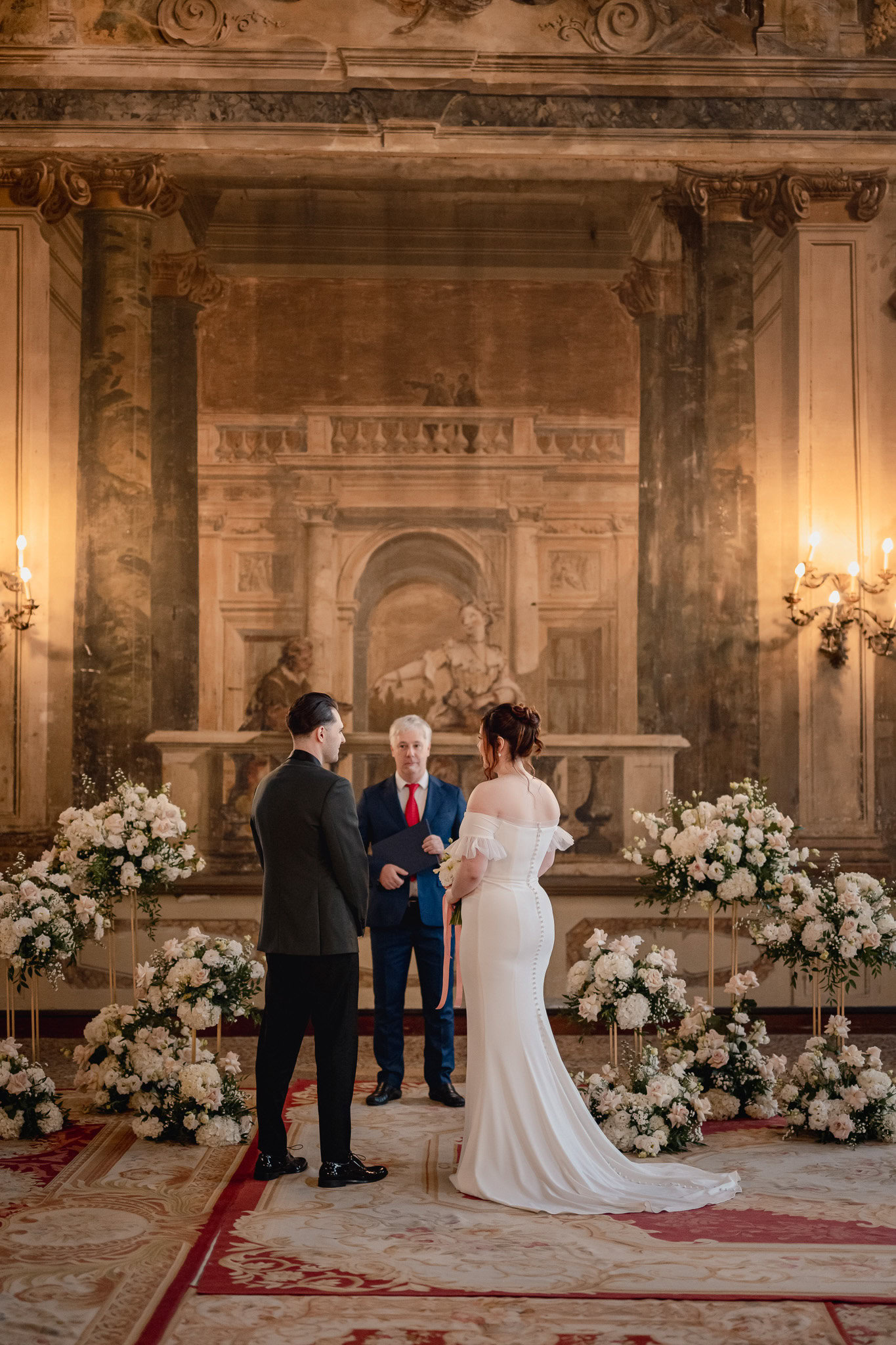 Elegant couple standing inside Ca’ Sagredo Palace with floral arrangements in the backgrou.