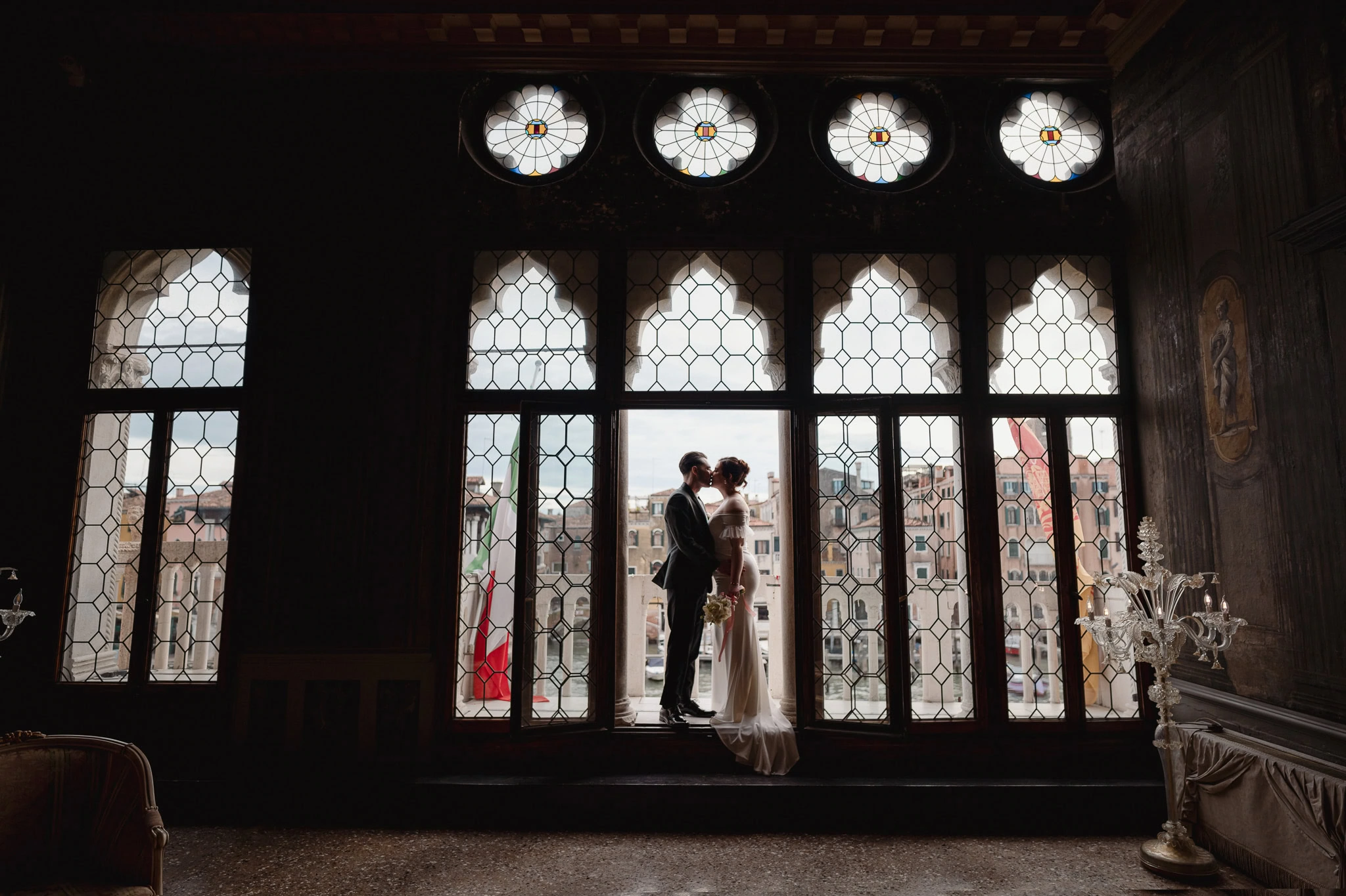 Silhouetted couple standing close inside Ca’ Sagredo Palace with ornate windows in Venice.