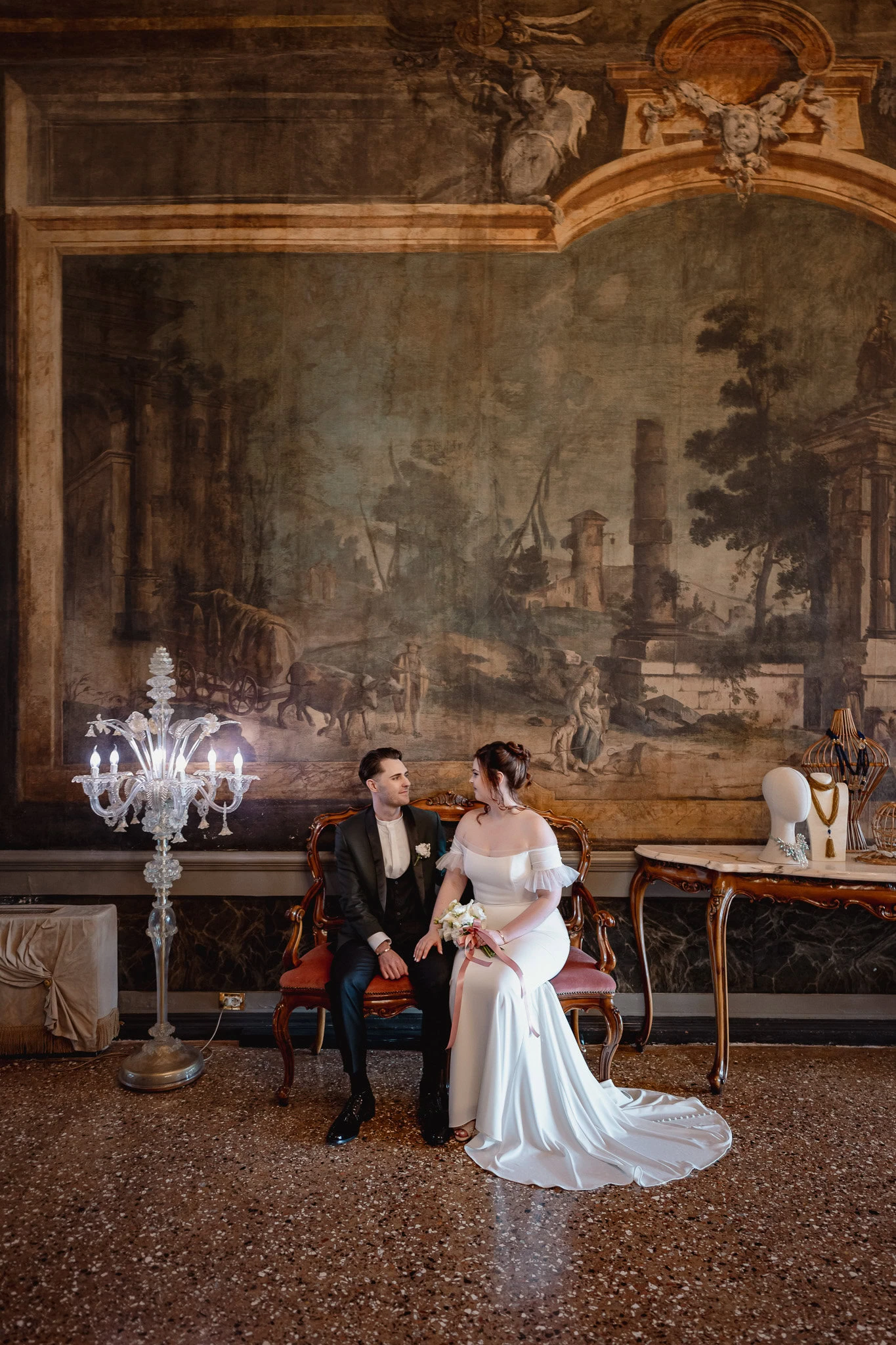 Elegant couple seated on a vintage sofa inside Ca’ Sagredo Palace, with a large classical.
