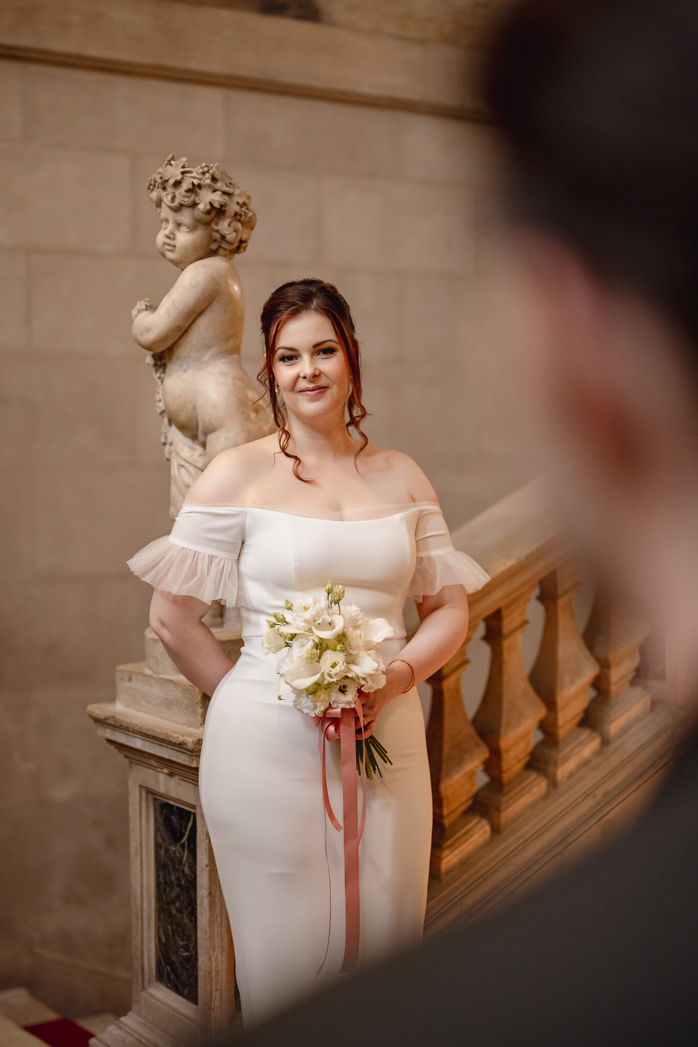 A woman in a white off-shoulder dress holding a bouquet at Ca’ Sagredo Palace.