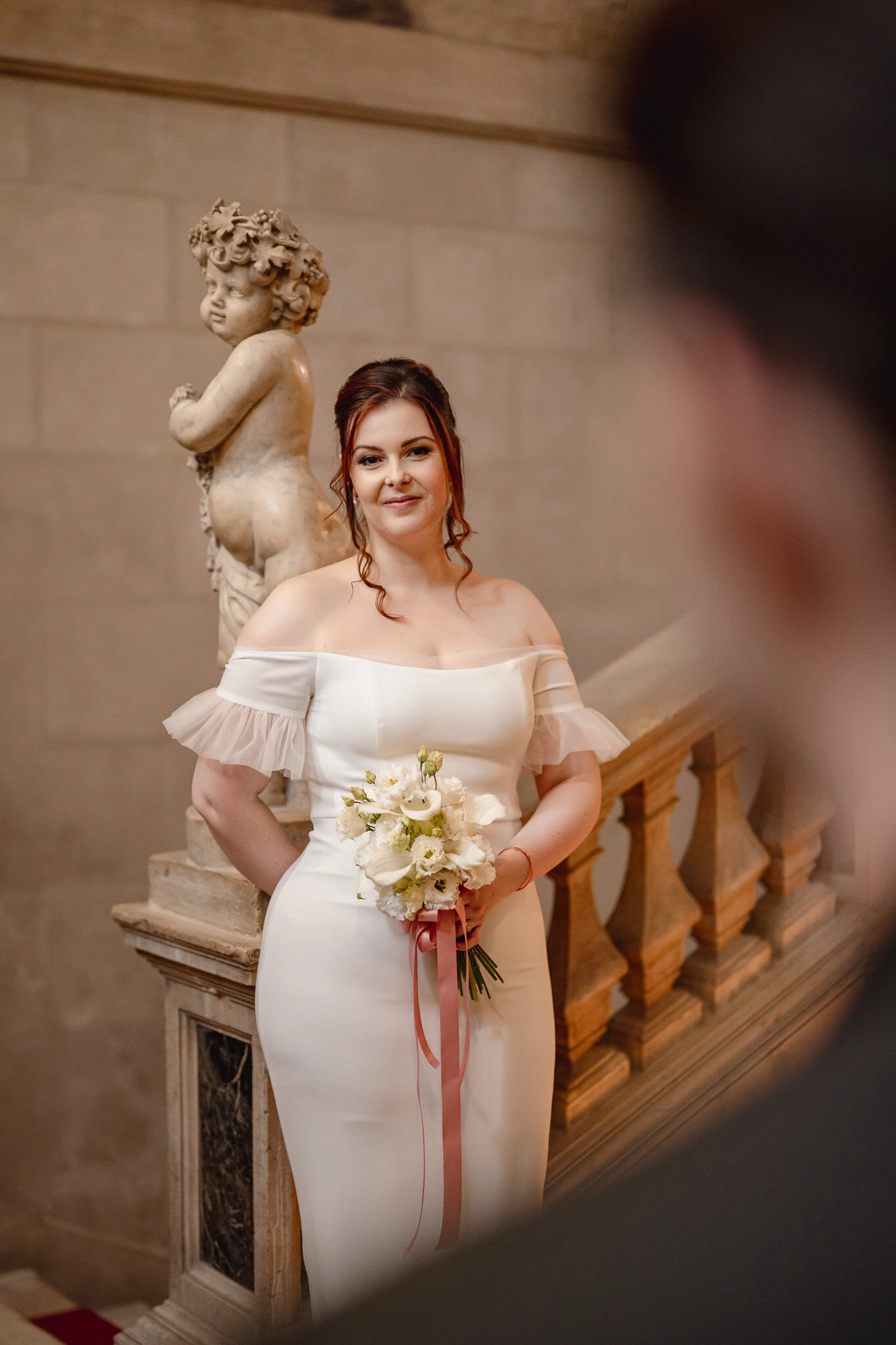 A woman in a white off-shoulder dress holding a bouquet at Ca’ Sagredo Palace.