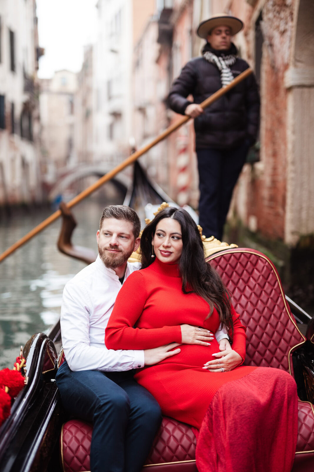 A smiling pregnant woman in a red dress sitting with her partner in a gondola in Venice.
