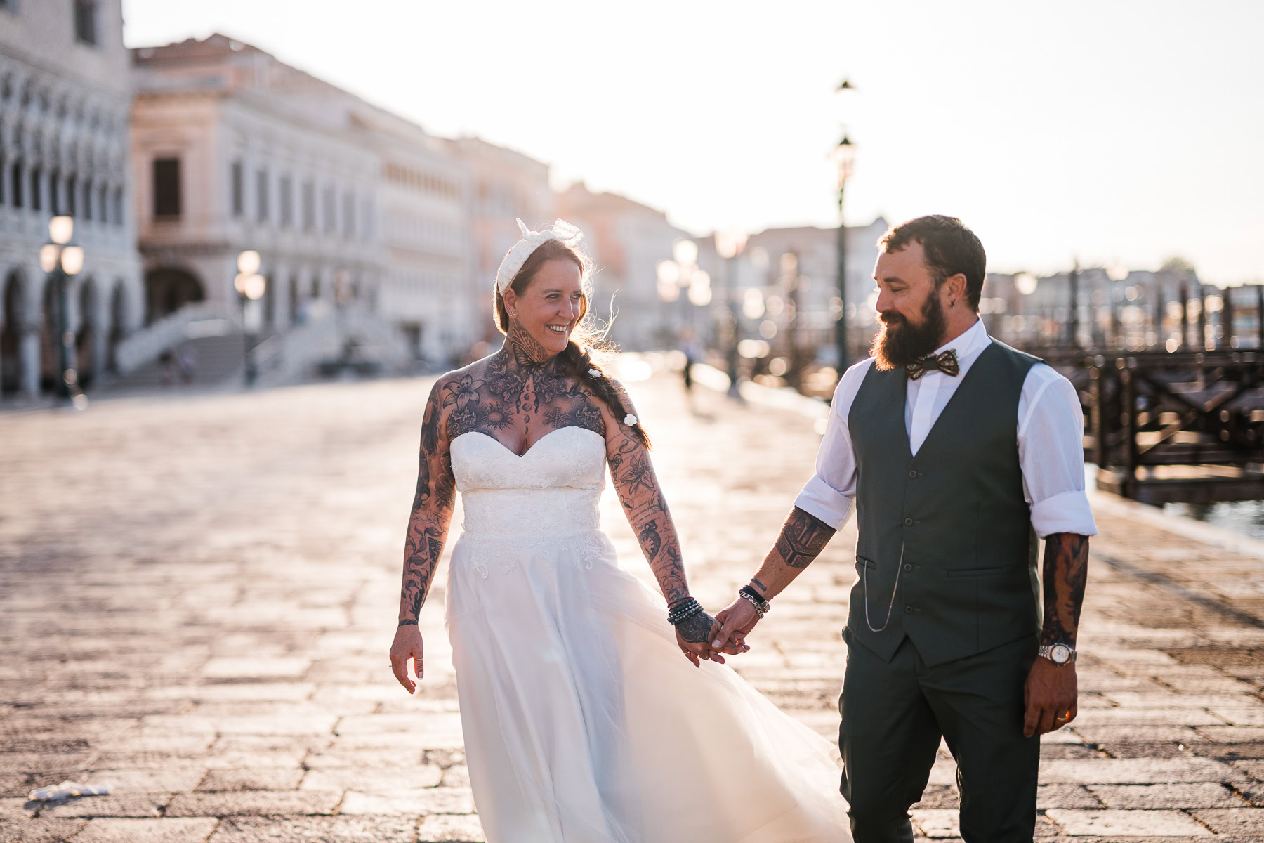 A woman and man holding hands, walking along a sunlit Venice street at dawn, with historic.