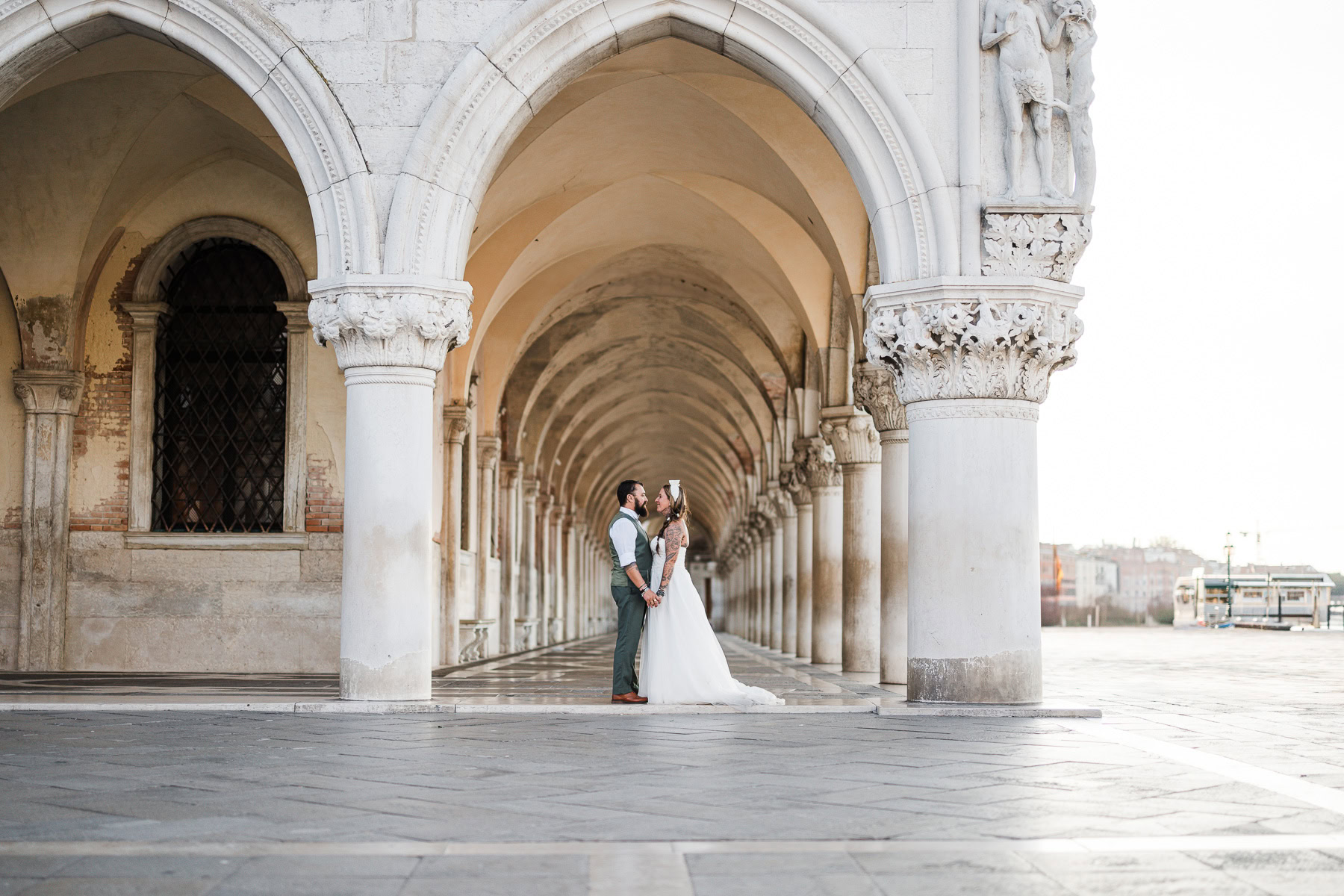 A woman in a wedding dress and a man in formal attire stand close under Venetian arches, illuminated by soft natural lig.