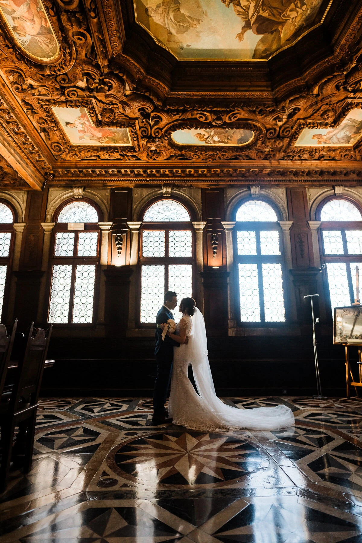 Couple standing inside Scuola Grande dei Carmini with ornate ceiling and large arched wind.