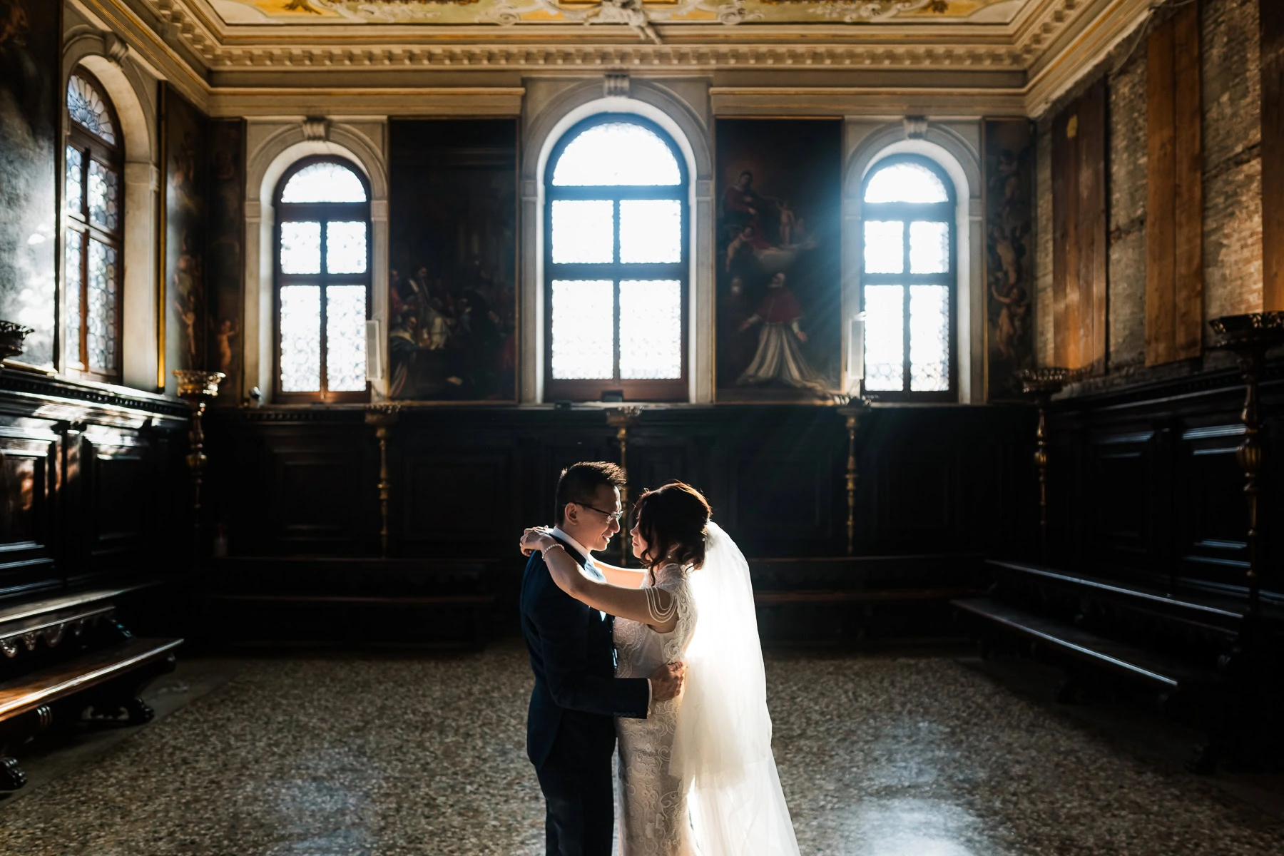 Couple embracing inside Scuola Grande dei Carmini with large arched windows in the backgro.