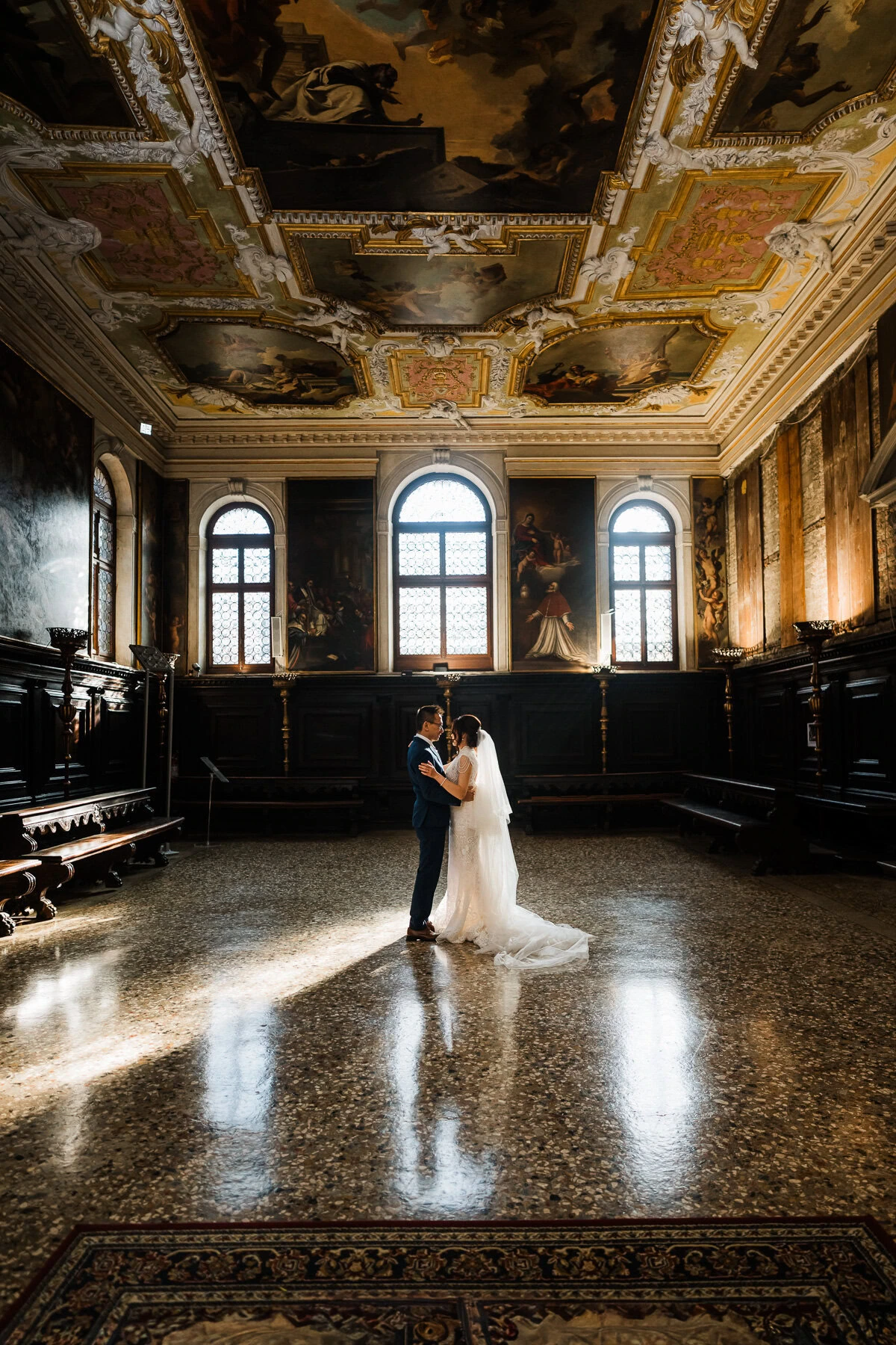 A woman and a man embrace in a grand, ornate room with high arched windows and a decorated.