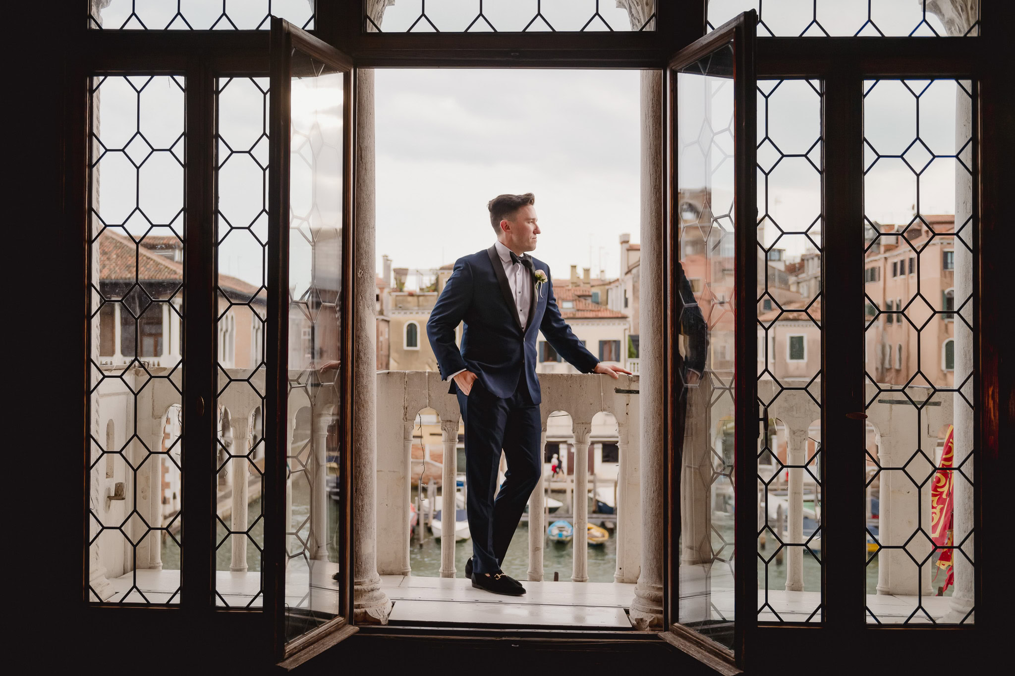 A man in a suit stands on a balcony inside a Venetian palace with open windows overlooking.