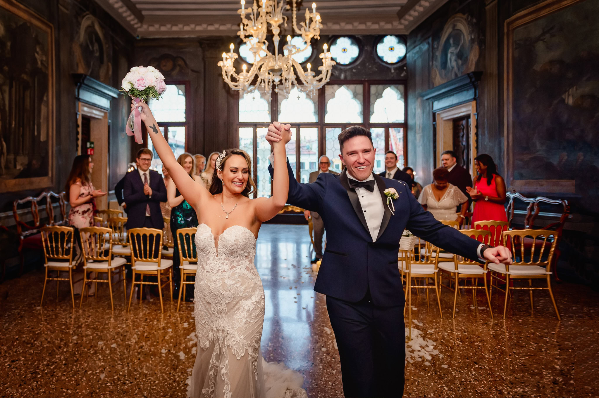 Newlywed couple celebrating in a Venetian palace with guests in the background.