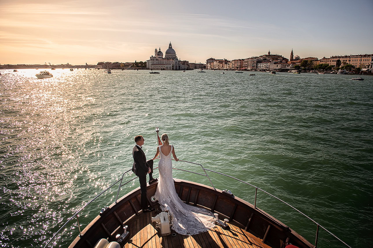 A couple shares an intimate moment on a boat in Venice during sunset, with historic cityscape and calm waters.