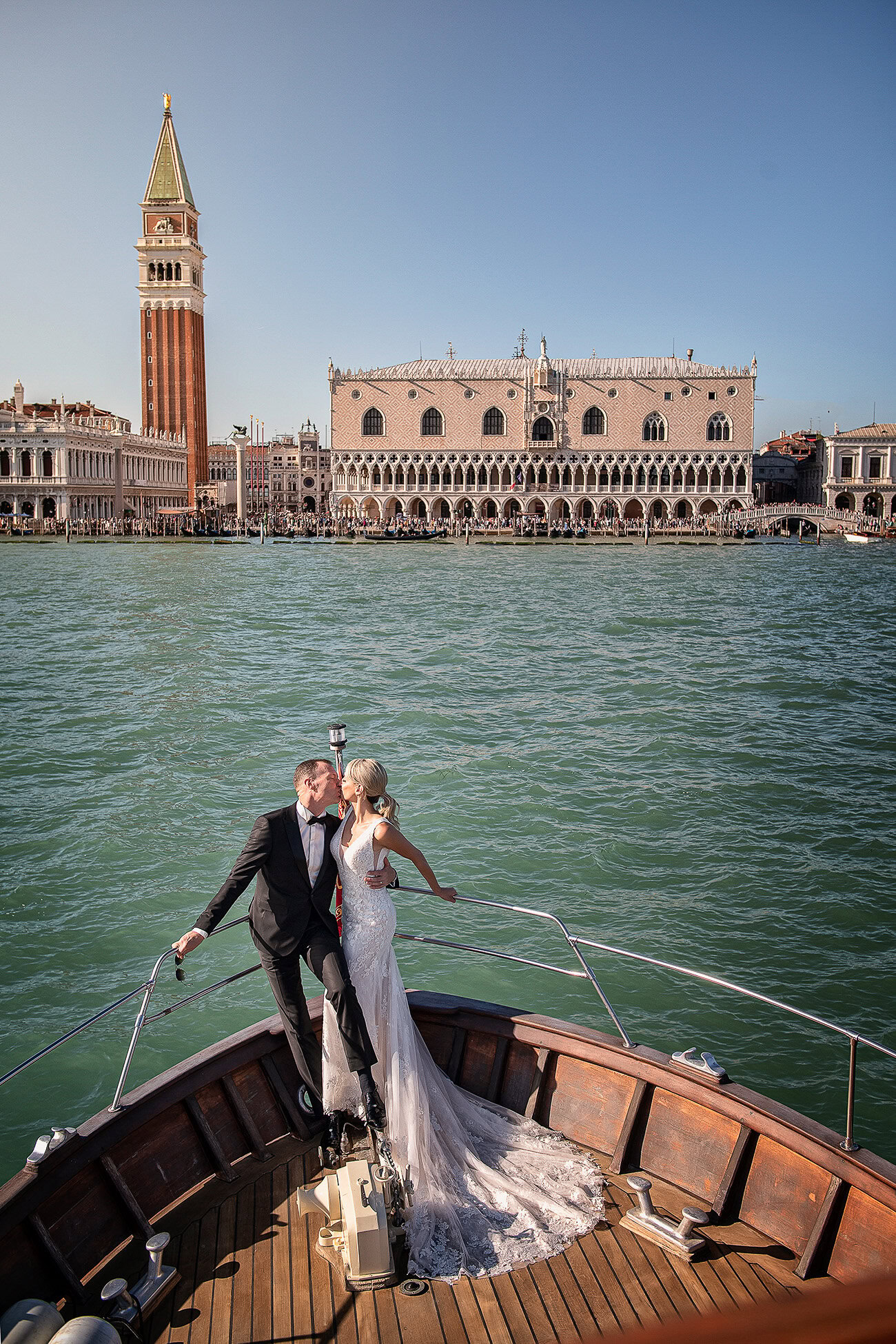 A couple in wedding attire standing on a boat in Venice with historic buildings and a bell tower in the background.