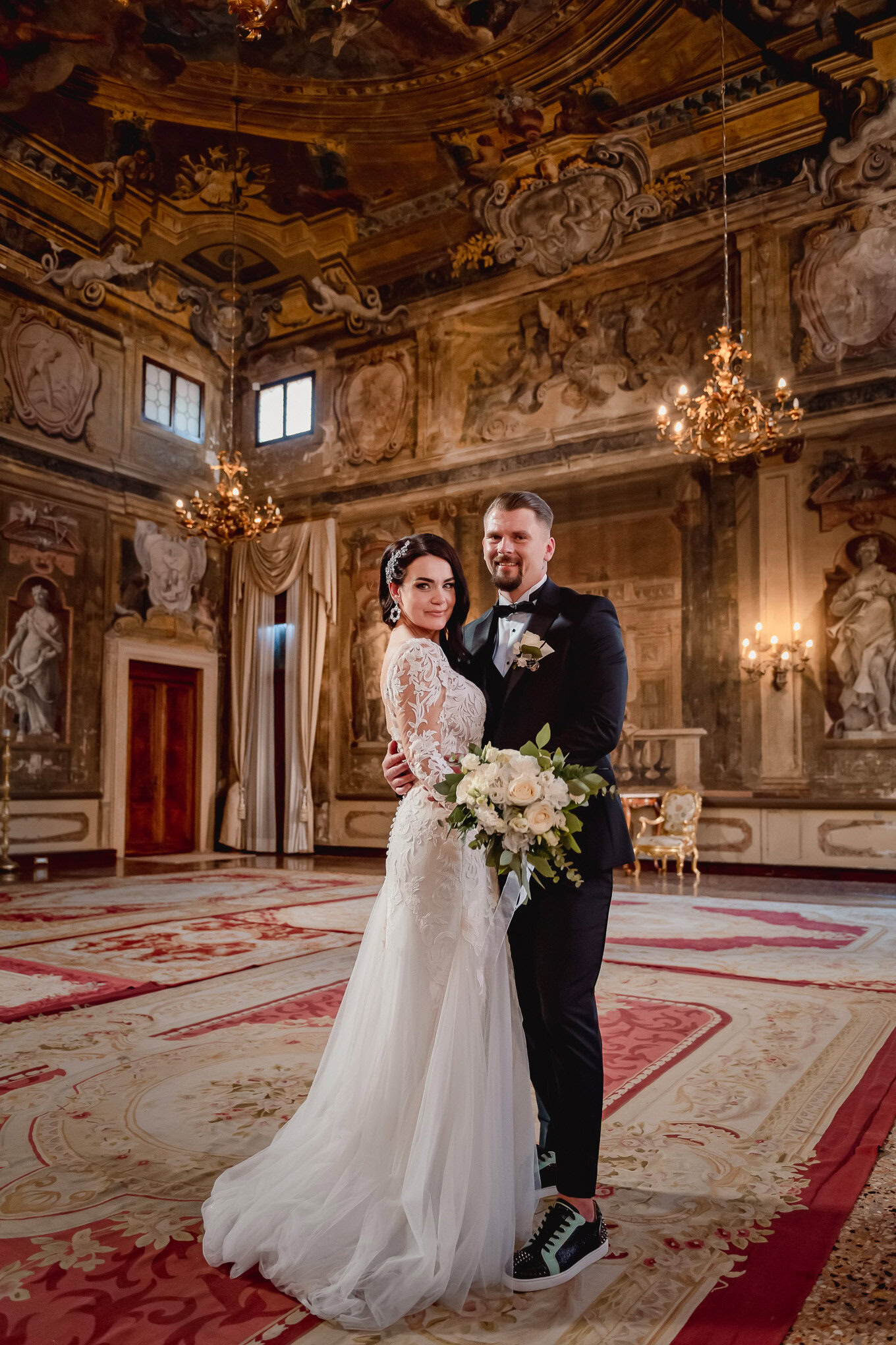 A detailed and intimate couple portrait inside an opulent, baroque-style hall with warm lighting and elaborate frescoes.