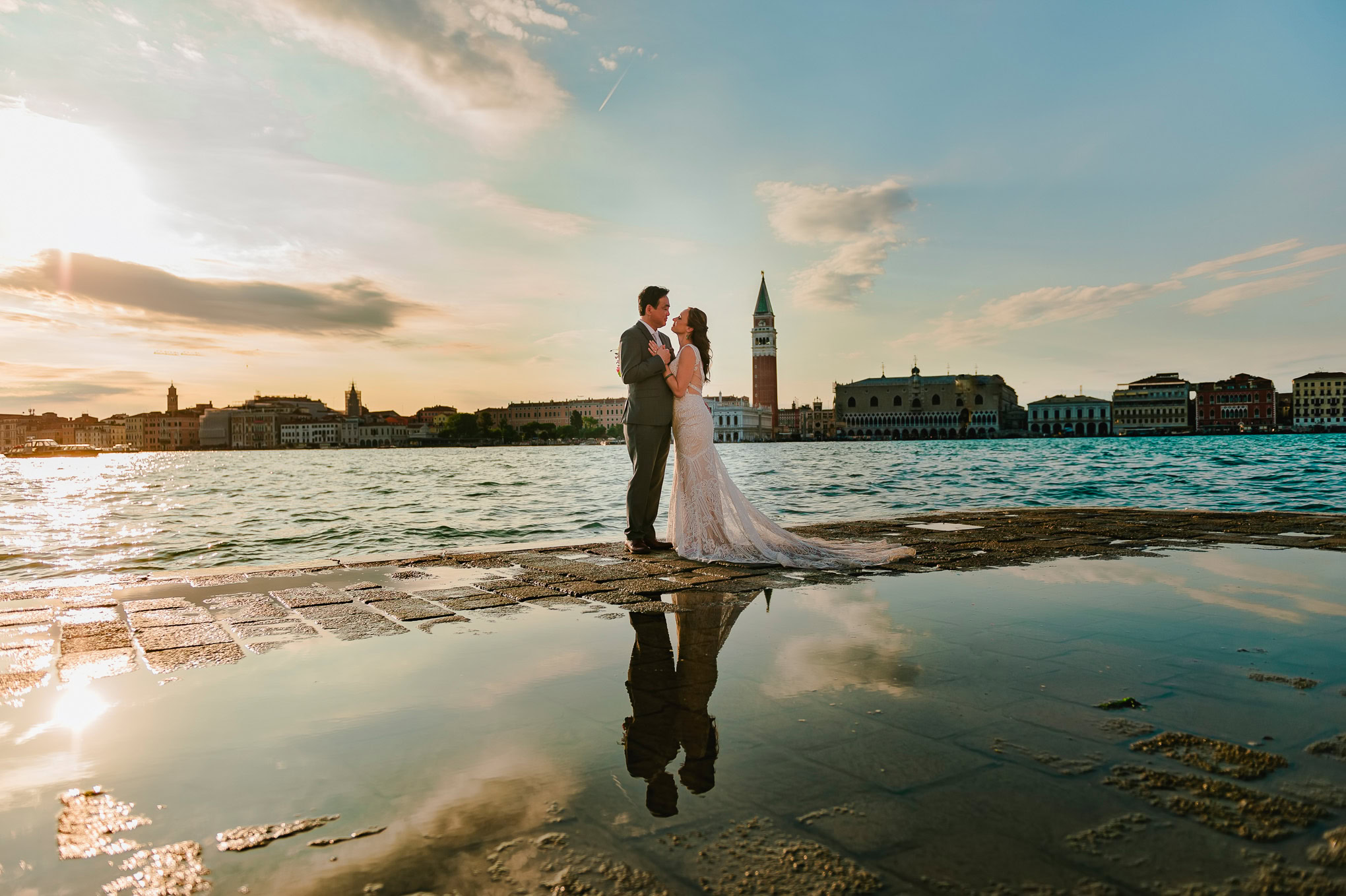 A couple shares an intimate moment by the water at sunset, with Venice’s skyline and the iconic bell tower in the background.