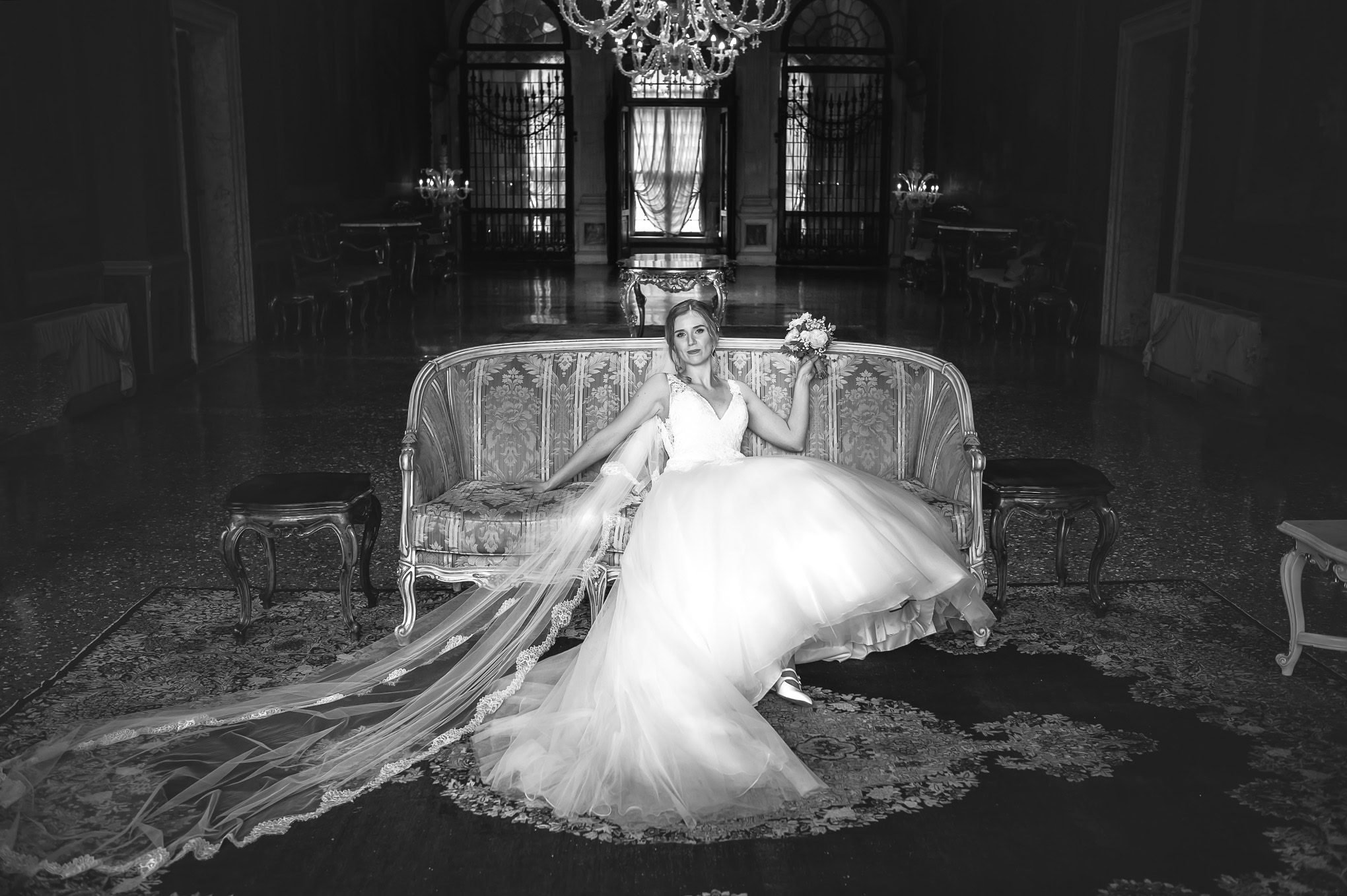 A woman in a flowing gown relaxes on an ornate sofa in a grand, dimly lit Venetian interior.