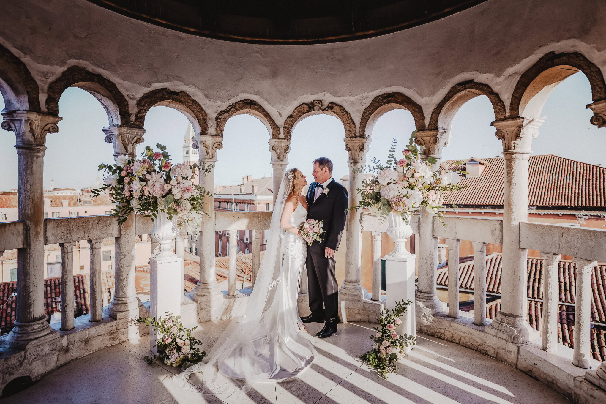 Intimate couple sharing a tender moment on a Venetian balcony bathed in soft natural light, surrounded by elegant floral.