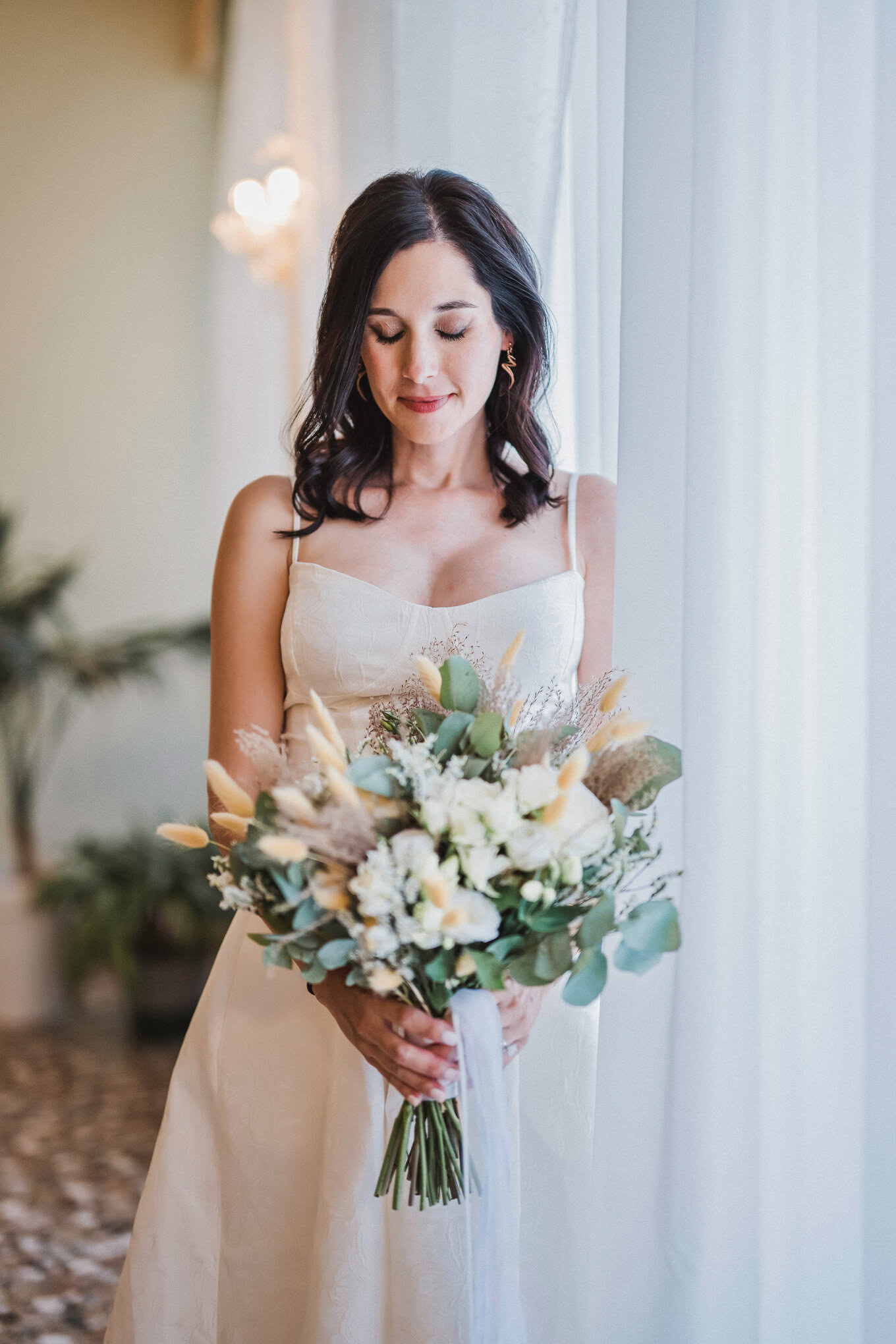 A woman in a white dress holding a bouquet of flowers, standing near a window with soft natural light.
