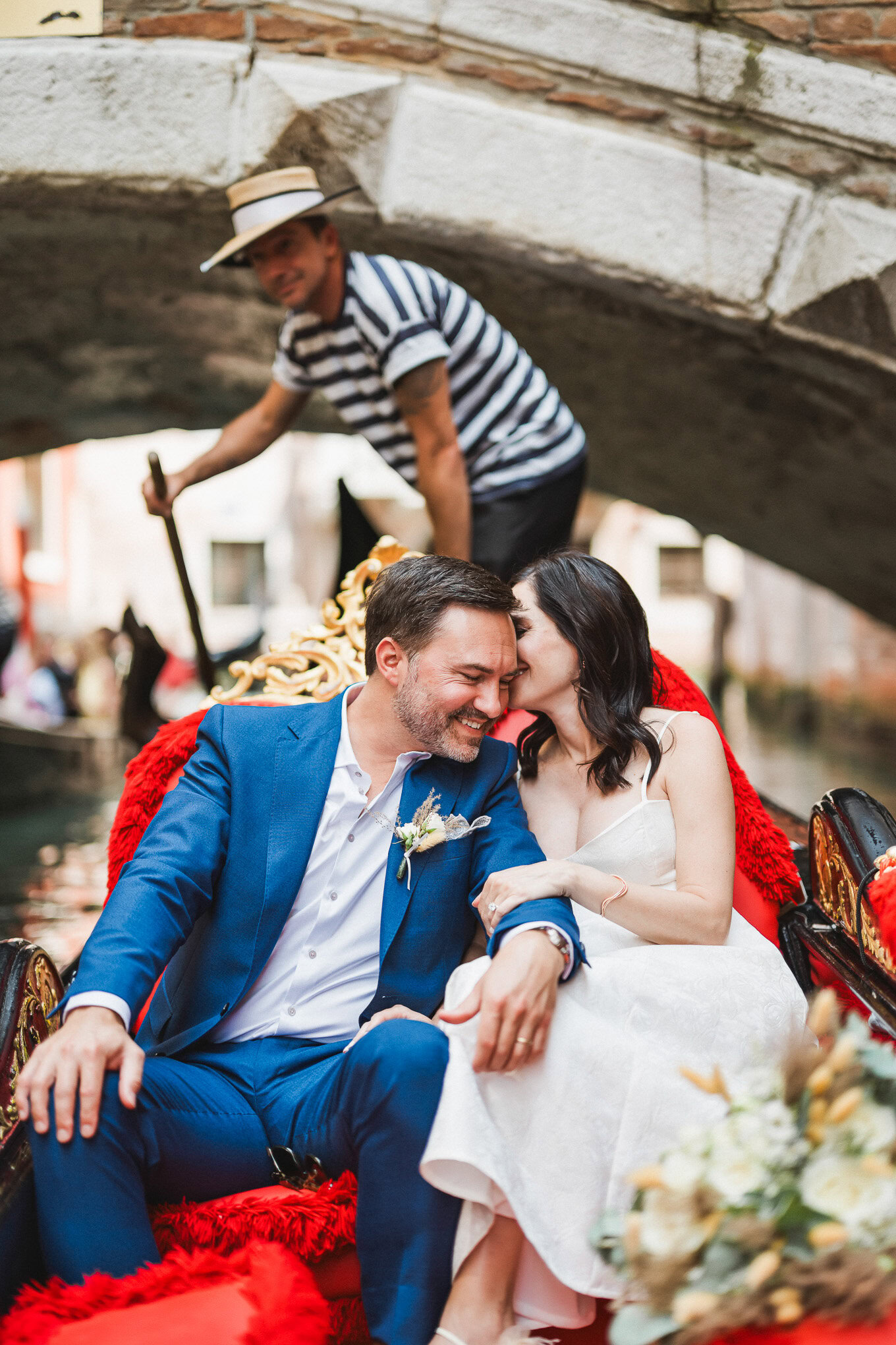 Intimate couple sharing a romantic moment on a gondola beneath a historic Venetian bridge, bathed in soft natural light.