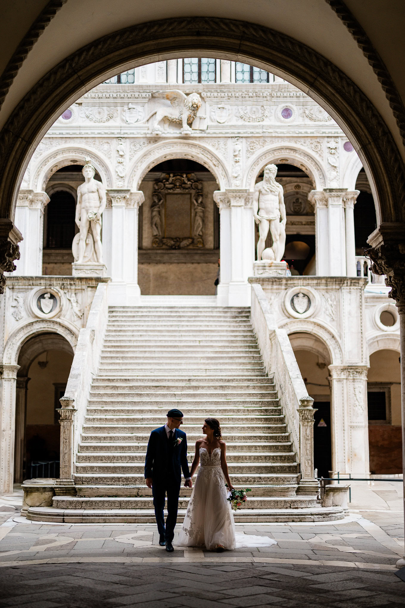A beautiful intimate couple walking hand in hand through a grand Venetian historic building, bathed in soft natural light.