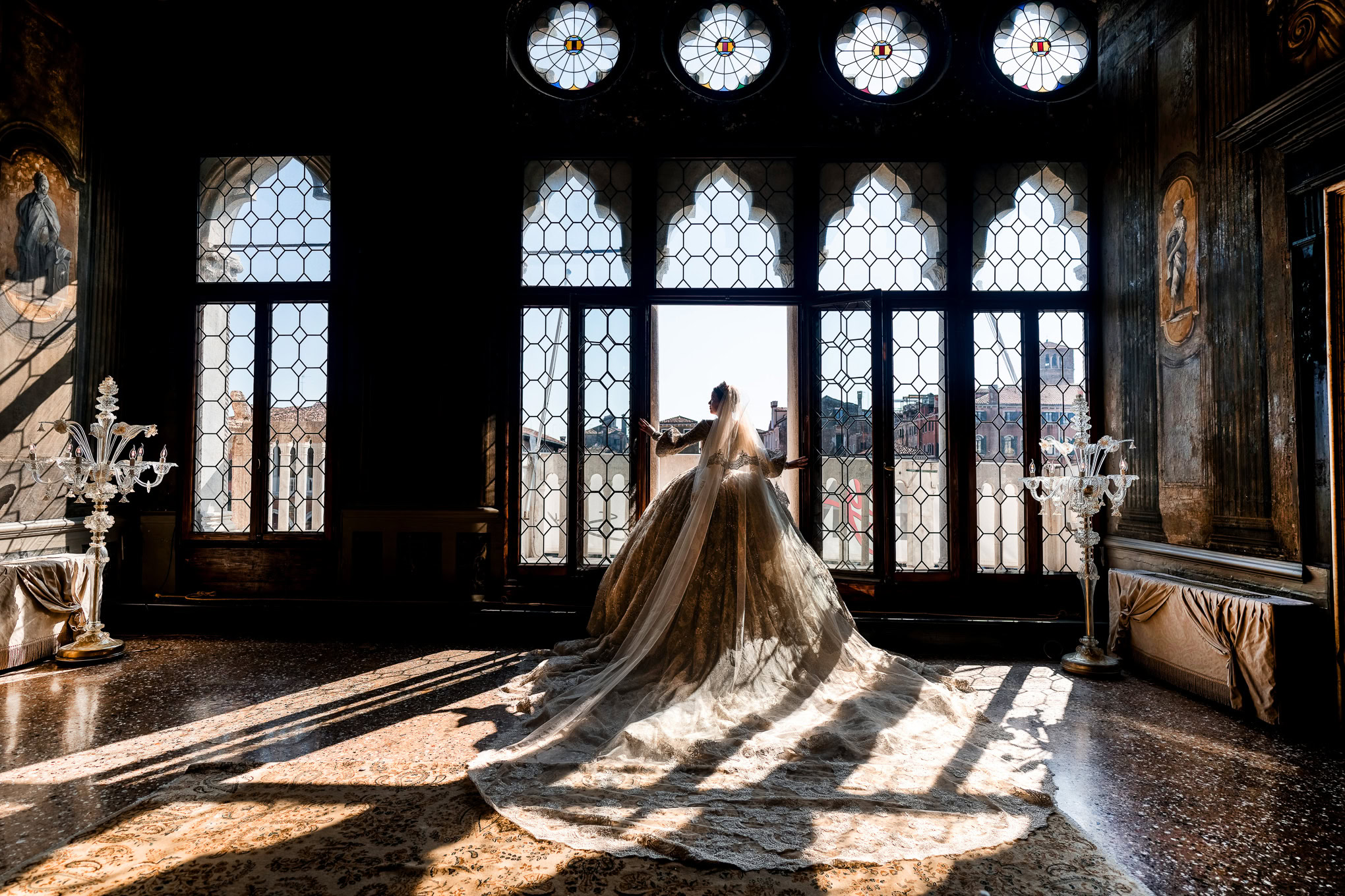 A woman in a gown stands in front of large, ornate windows, bathed in warm sunlight.