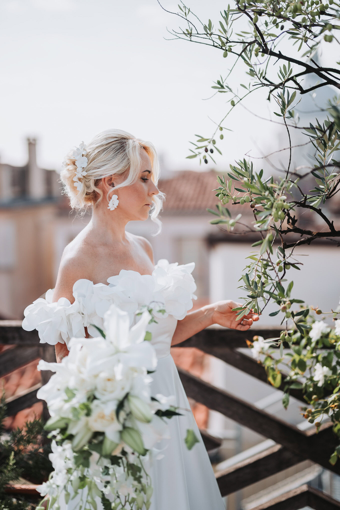 A woman in a white wedding dress stands on a Venetian balcony, surrounded by greenery, with soft natural light highlight.