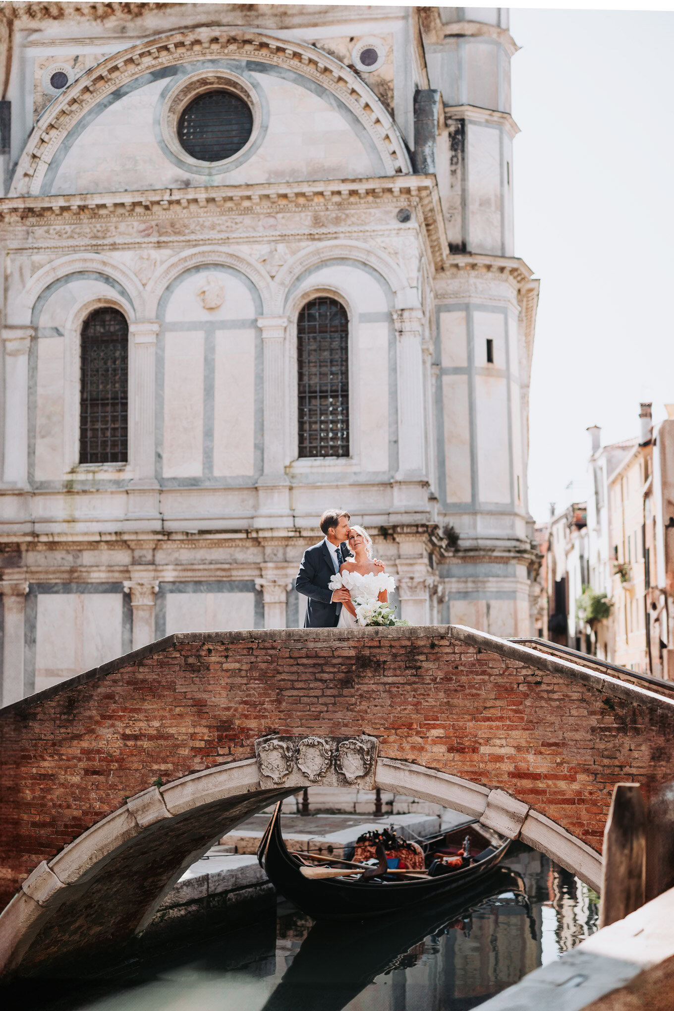 A couple sharing an intimate moment on a small bridge in Venice, bathed in soft daylight with historic architecture.