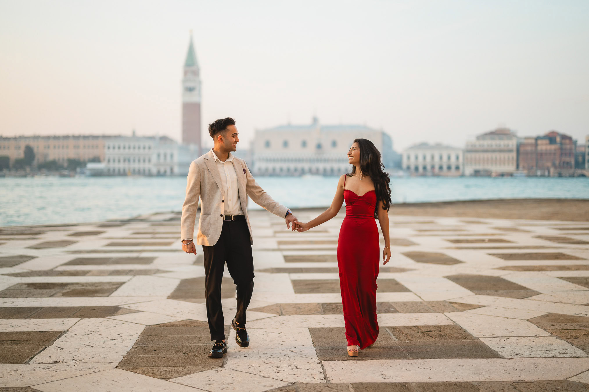A man and woman holding hands and walking along a waterfront promenade in Venice, with historic buil.