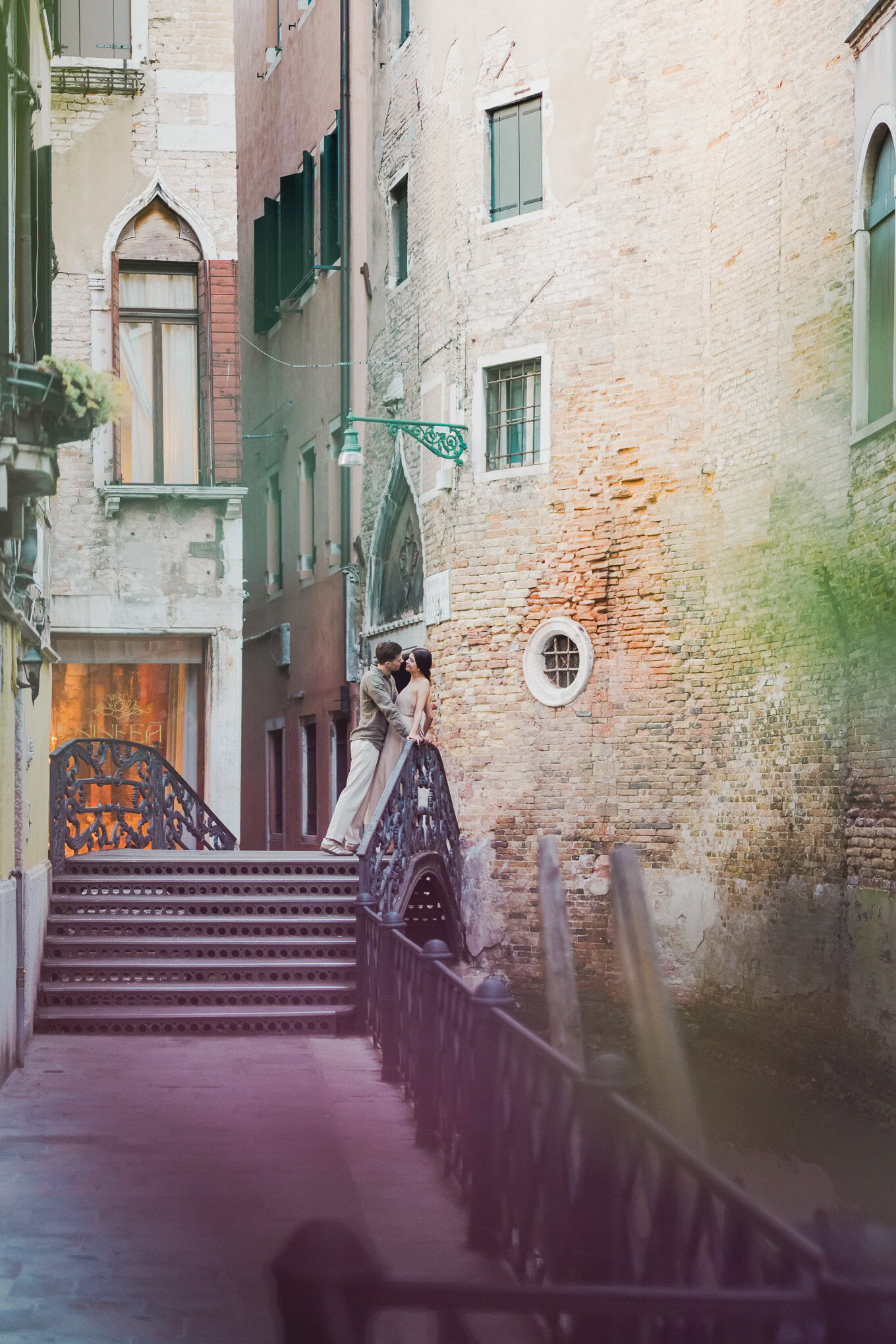 A couple shares a tender moment on a narrow, sunlit Venetian canal staircase, surrounded by weathere.