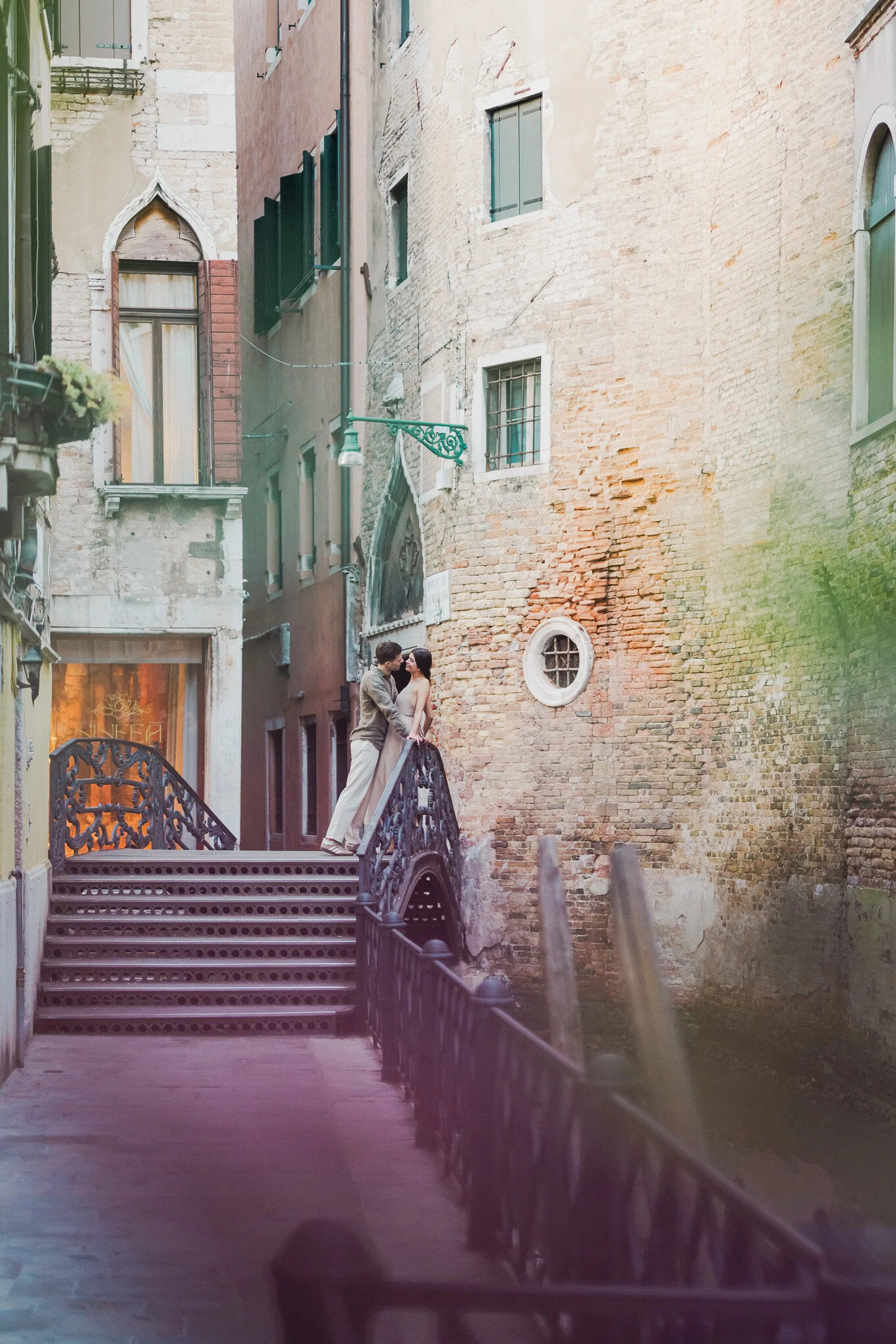 A couple shares a tender moment on a narrow, sunlit Venetian canal staircase, surrounded by weathere.