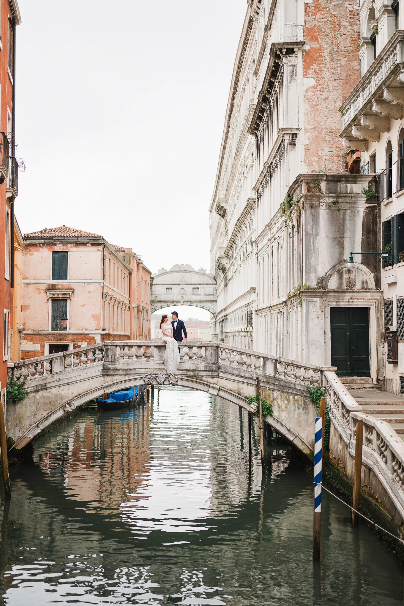 A couple shares a tender moment on a small bridge over a canal in Venice, bathed in soft natural lig.
