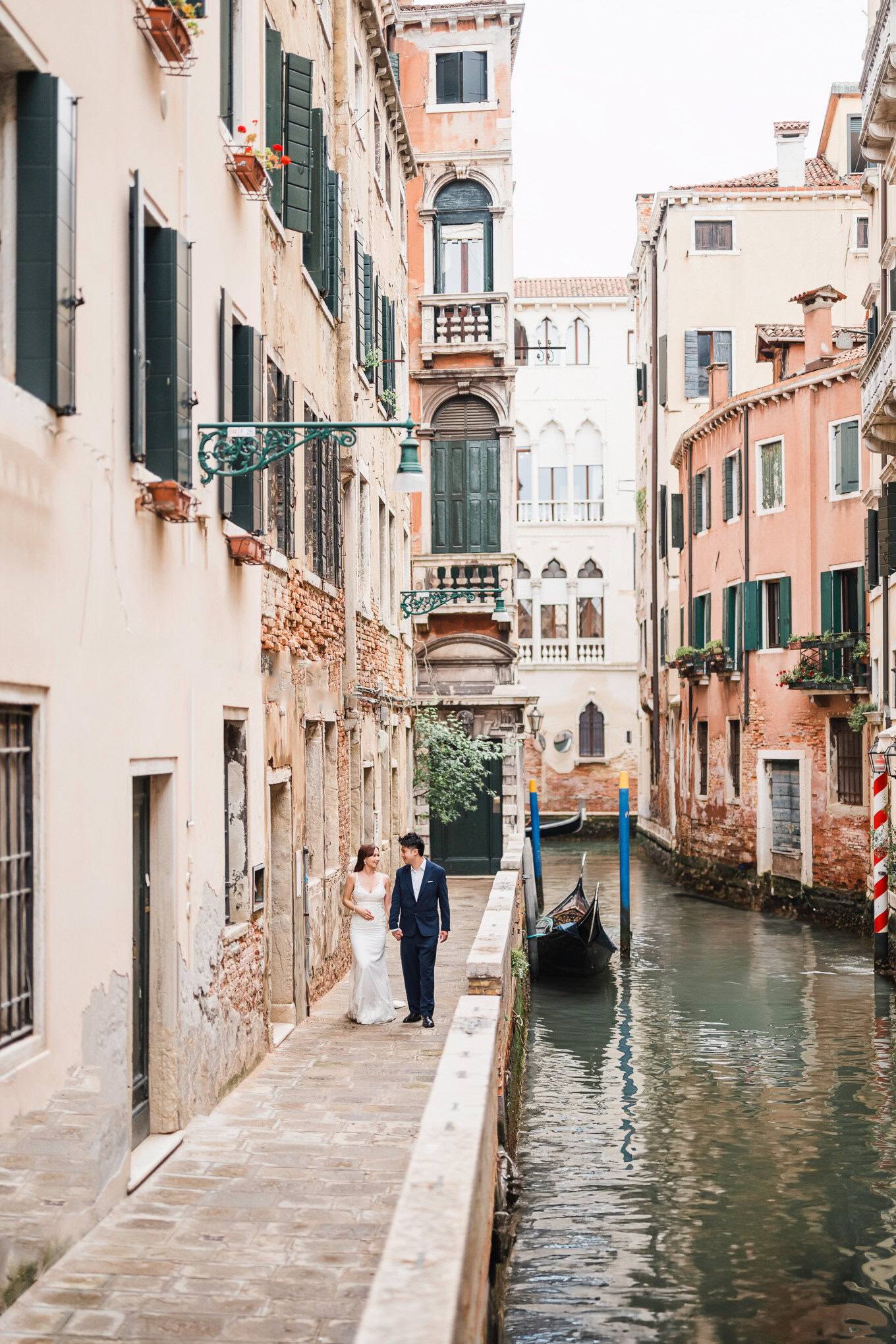 A couple in wedding attire walking along a narrow canal in Venice, bathed in soft daylight, evoking.
