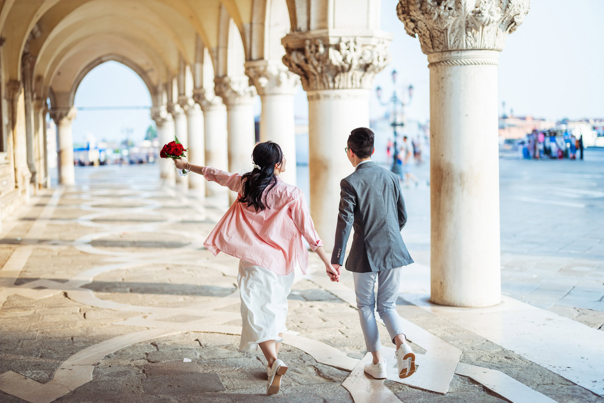 A woman and man holding hands and walking under historic arches on a sunny day, evoking romance and.