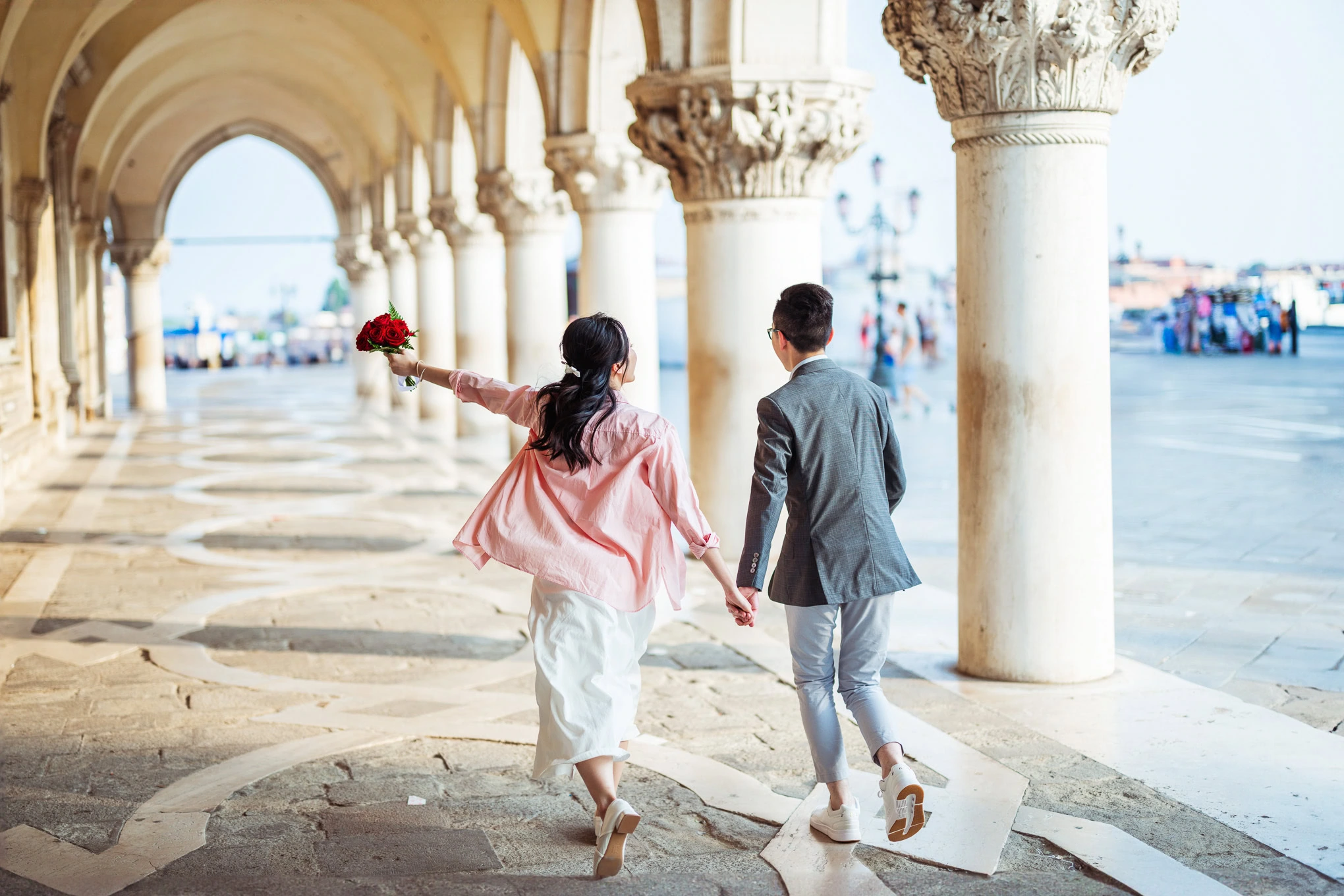 A woman and man holding hands and walking under historic arches on a sunny day, evoking romance and.