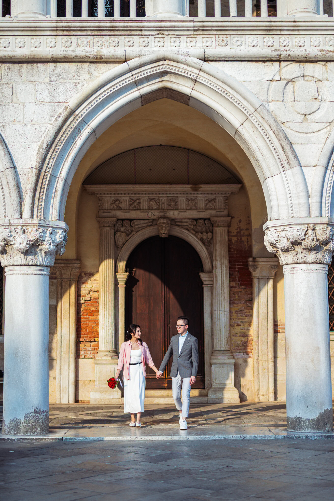 A couple holding hands and walking through an ancient stone archway at sunset, evoking a sense of ro.