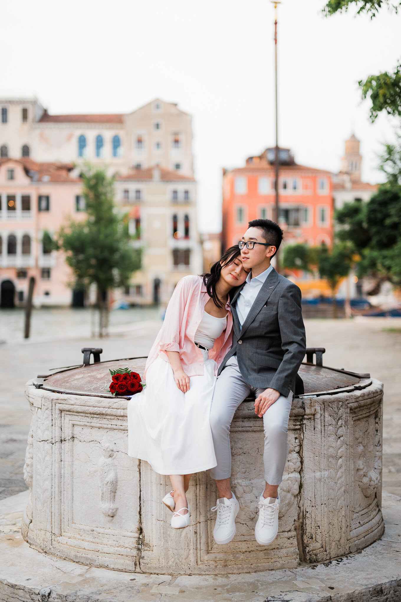 A woman resting her head on a man's shoulder while sitting on a stone fountain in a charming city sq.