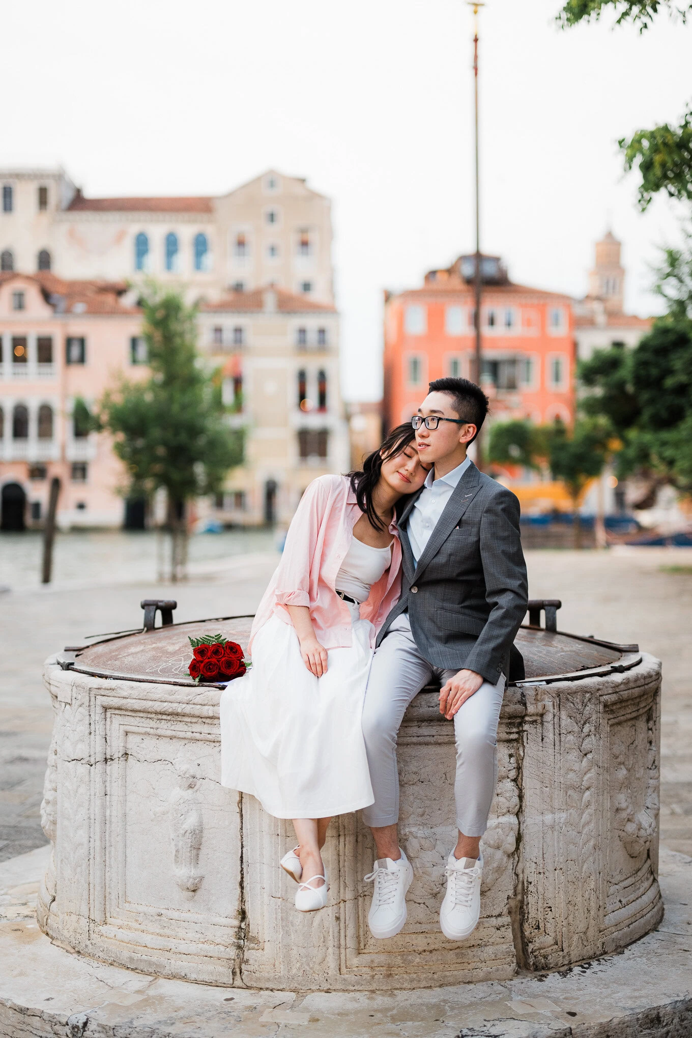 A woman resting her head on a man's shoulder while sitting on a stone fountain in a charming city sq.