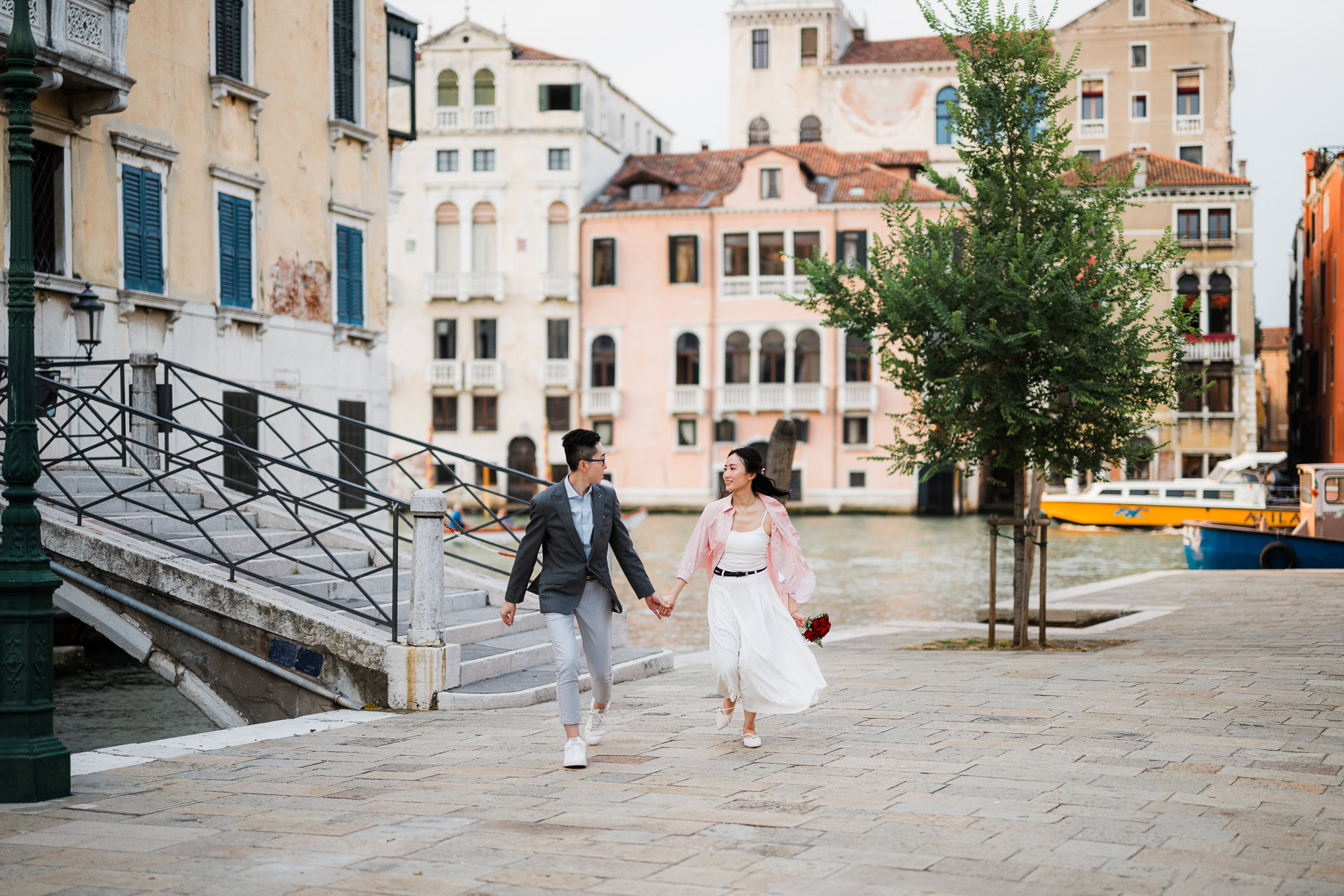 A couple holding hands and walking along a canal in Venice, surrounded by historic buildings and a p.