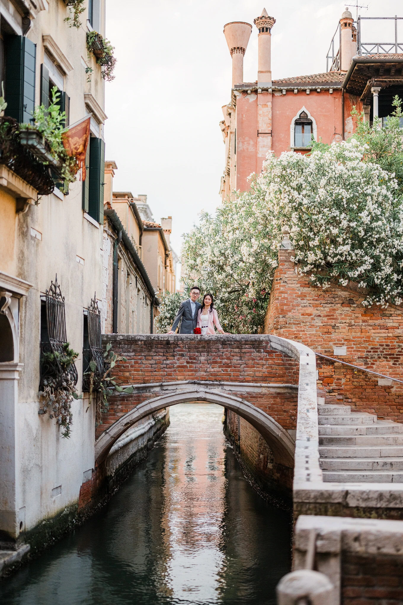 A couple stands on a small bridge over a narrow canal in Venice, surrounded by blooming trees and hi.