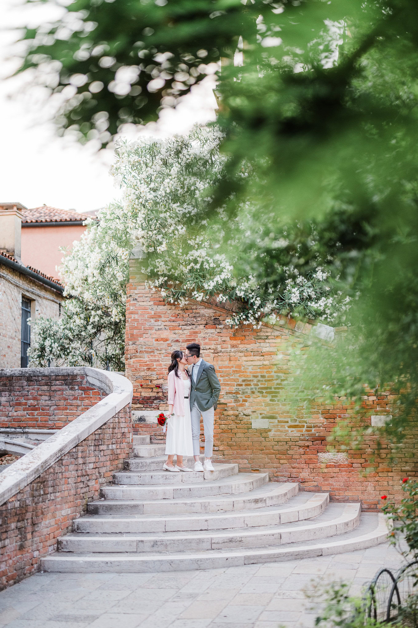 A couple standing on a Venetian staircase surrounded by lush greenery and blooming trees, capturing a romantic atmospher.