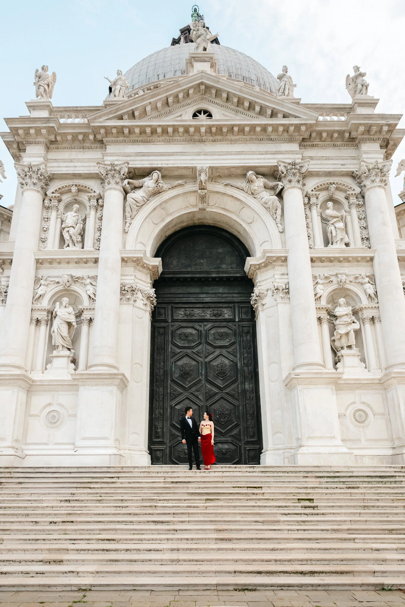 A woman in a red dress and a man in formal attire stand together on the steps of an ornate, historic.