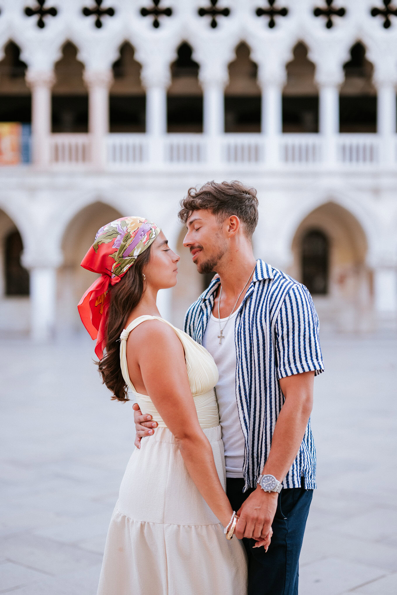 A woman and man share an intimate moment in a historic, sunlit courtyard, their faces close with ten.