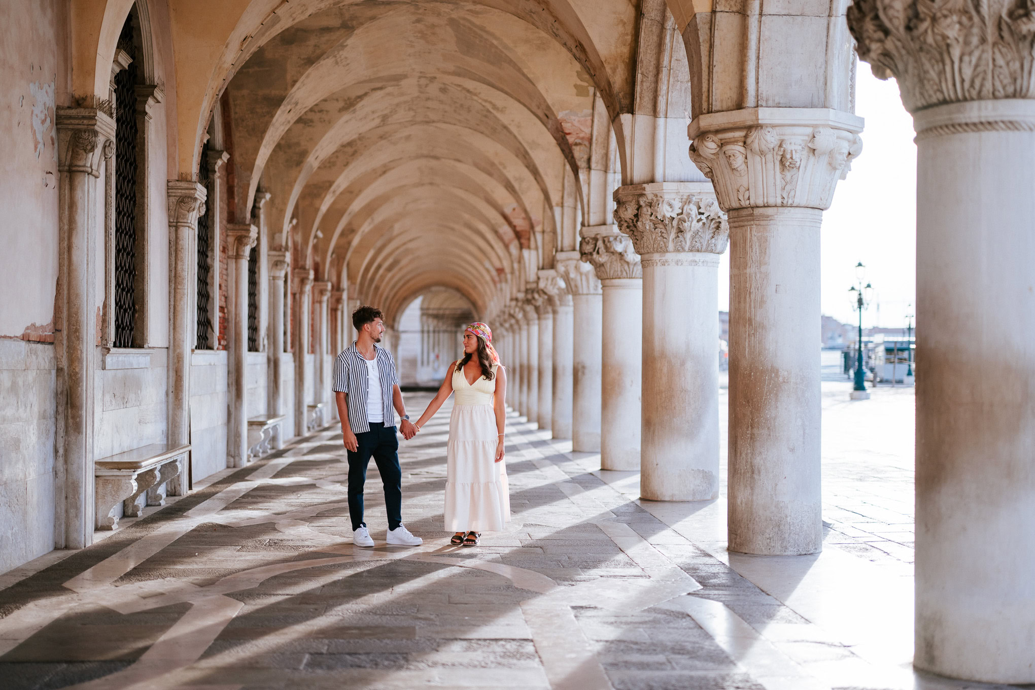A serene moment of a couple holding hands beneath historic arches bathed in soft sunlight, evoking i.