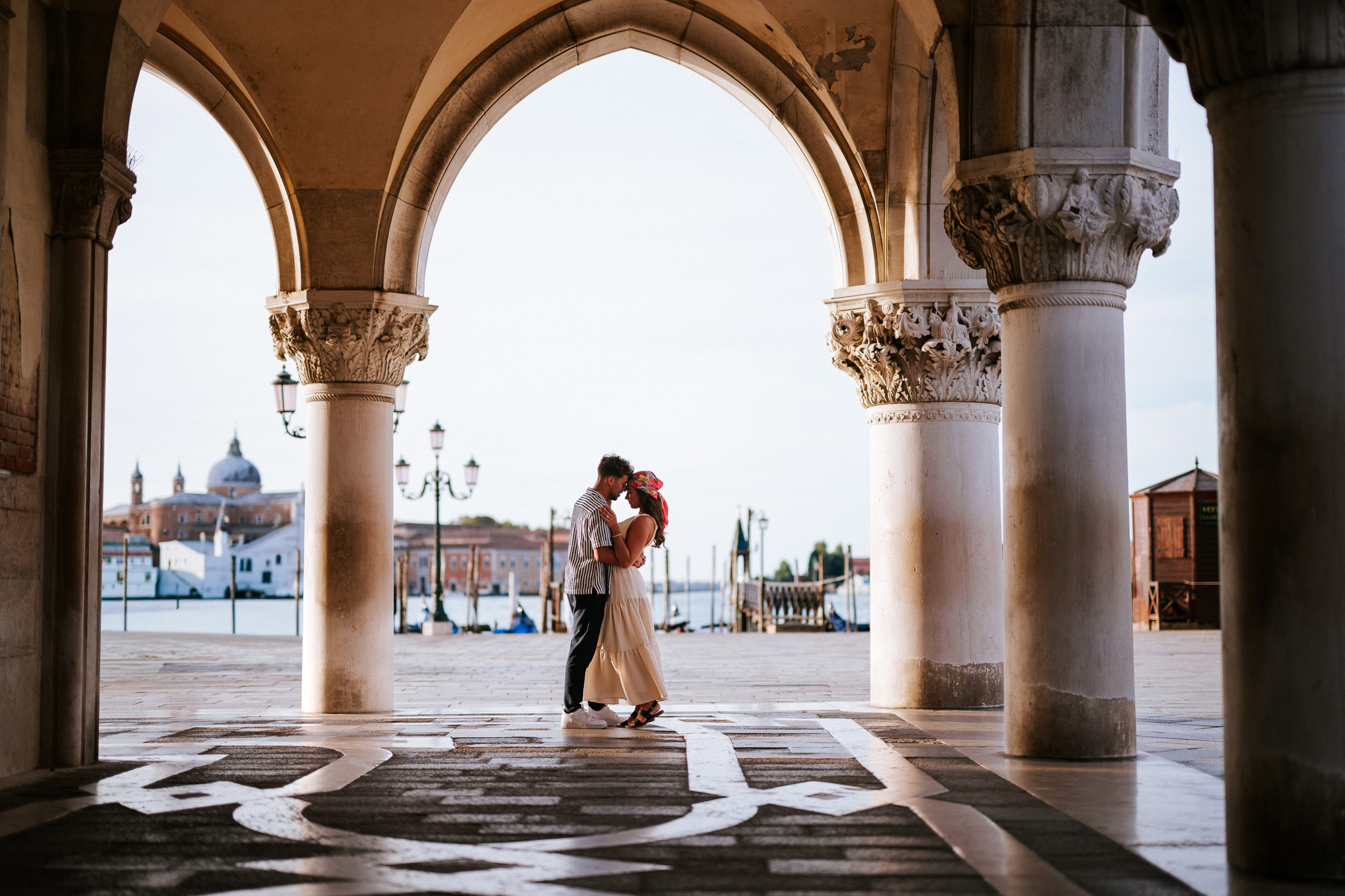 A couple sharing an intimate moment under historic arches by the water at sunset, evoking romance an.