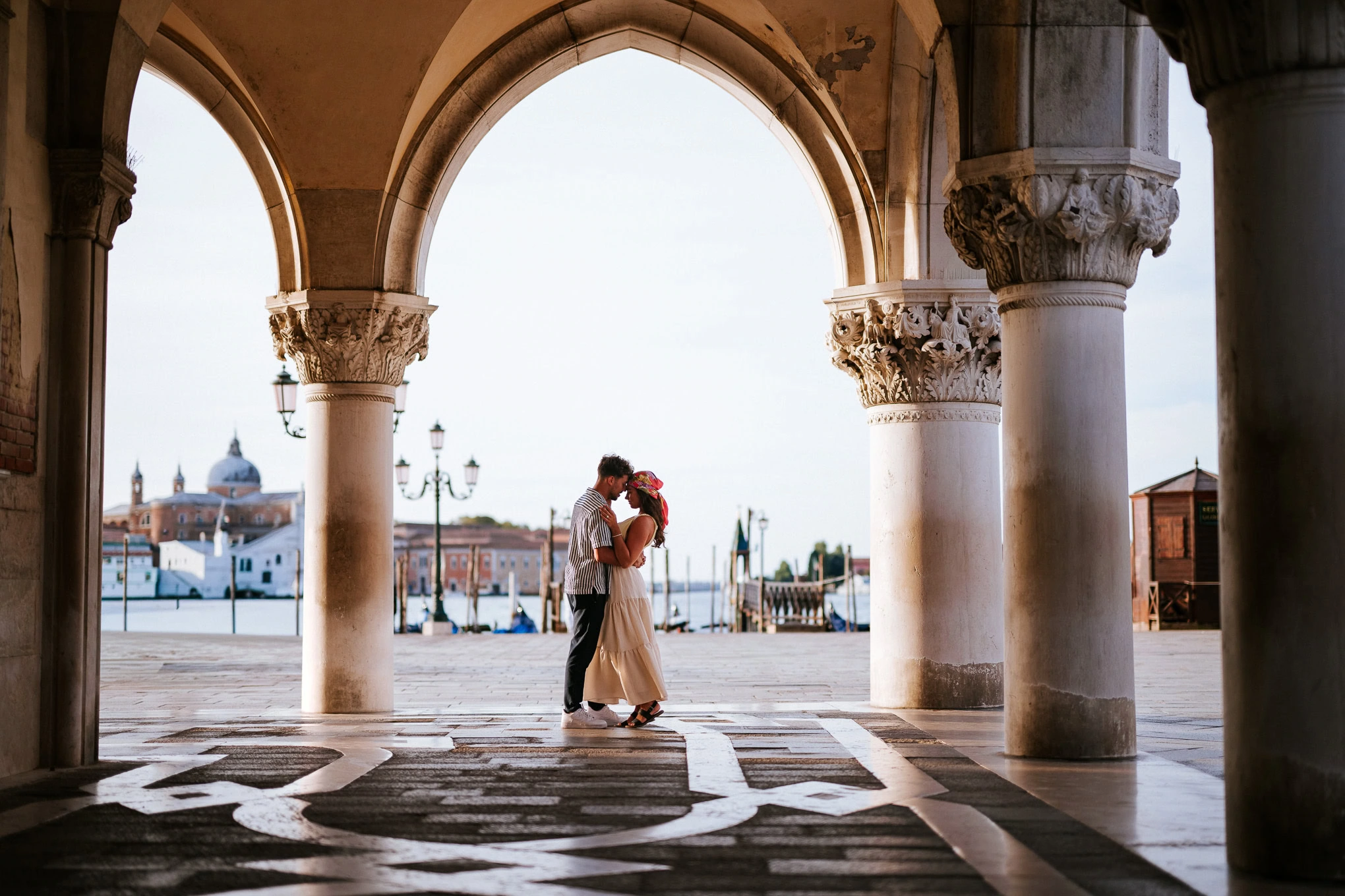 A couple sharing an intimate moment under historic arches by the water at sunset, evoking romance an.