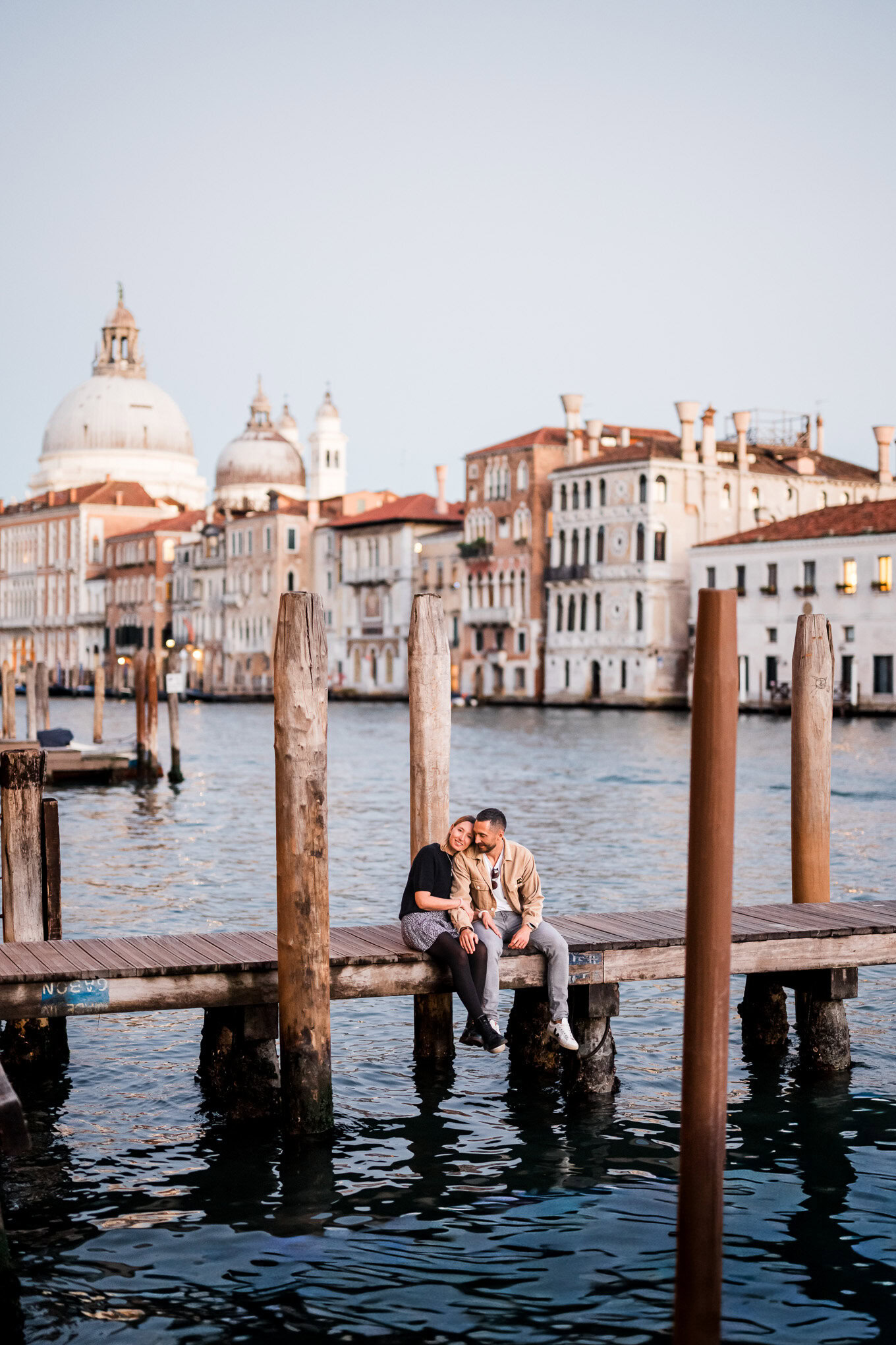 A couple sharing an intimate moment on a wooden dock in Venice at sunset, with historic buildings an.